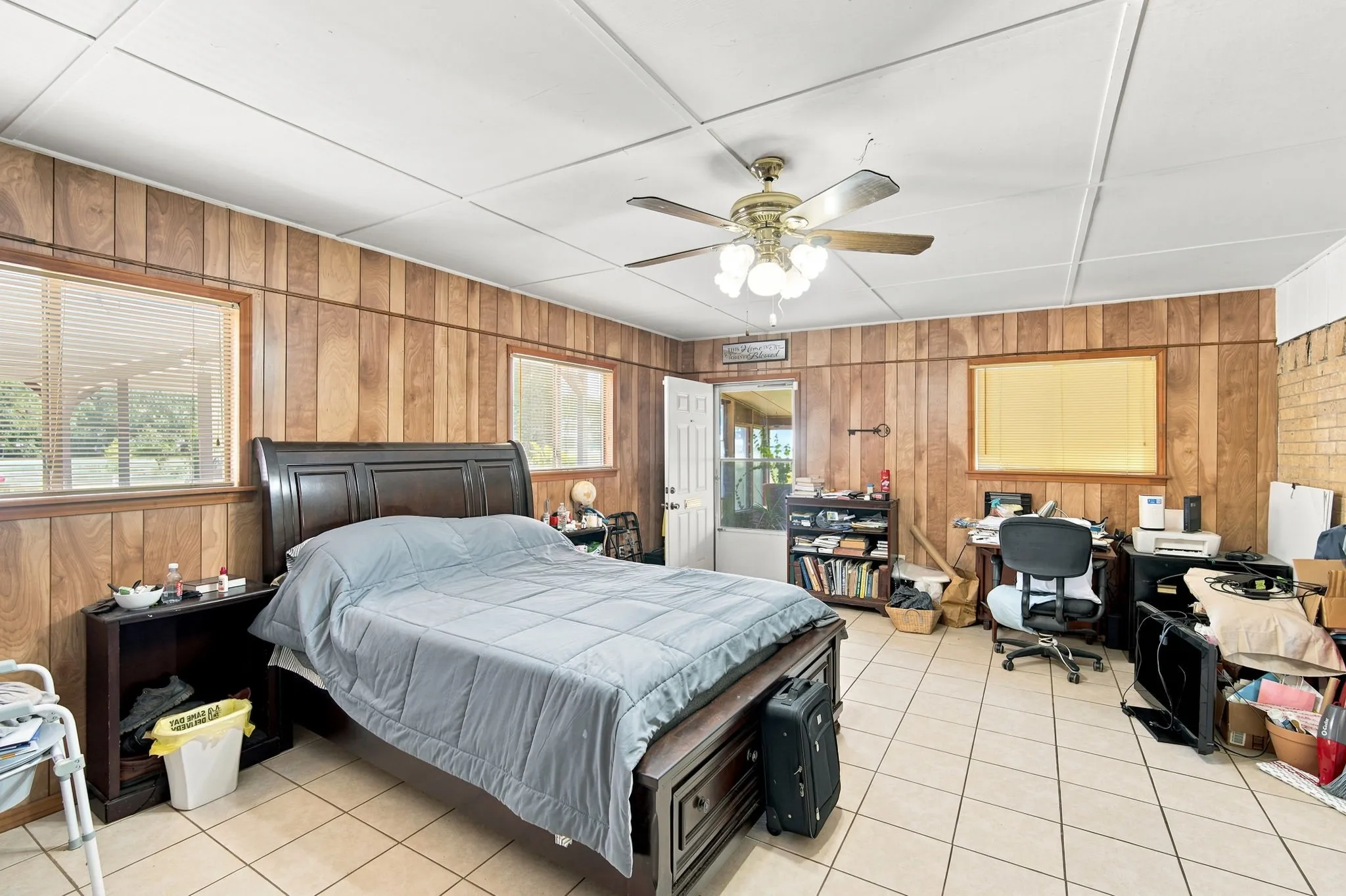 Spacious room featuring wood paneling, tiled flooring, multiple windows with blinds, a ceiling fan with integrated lighting, and a white paneled door