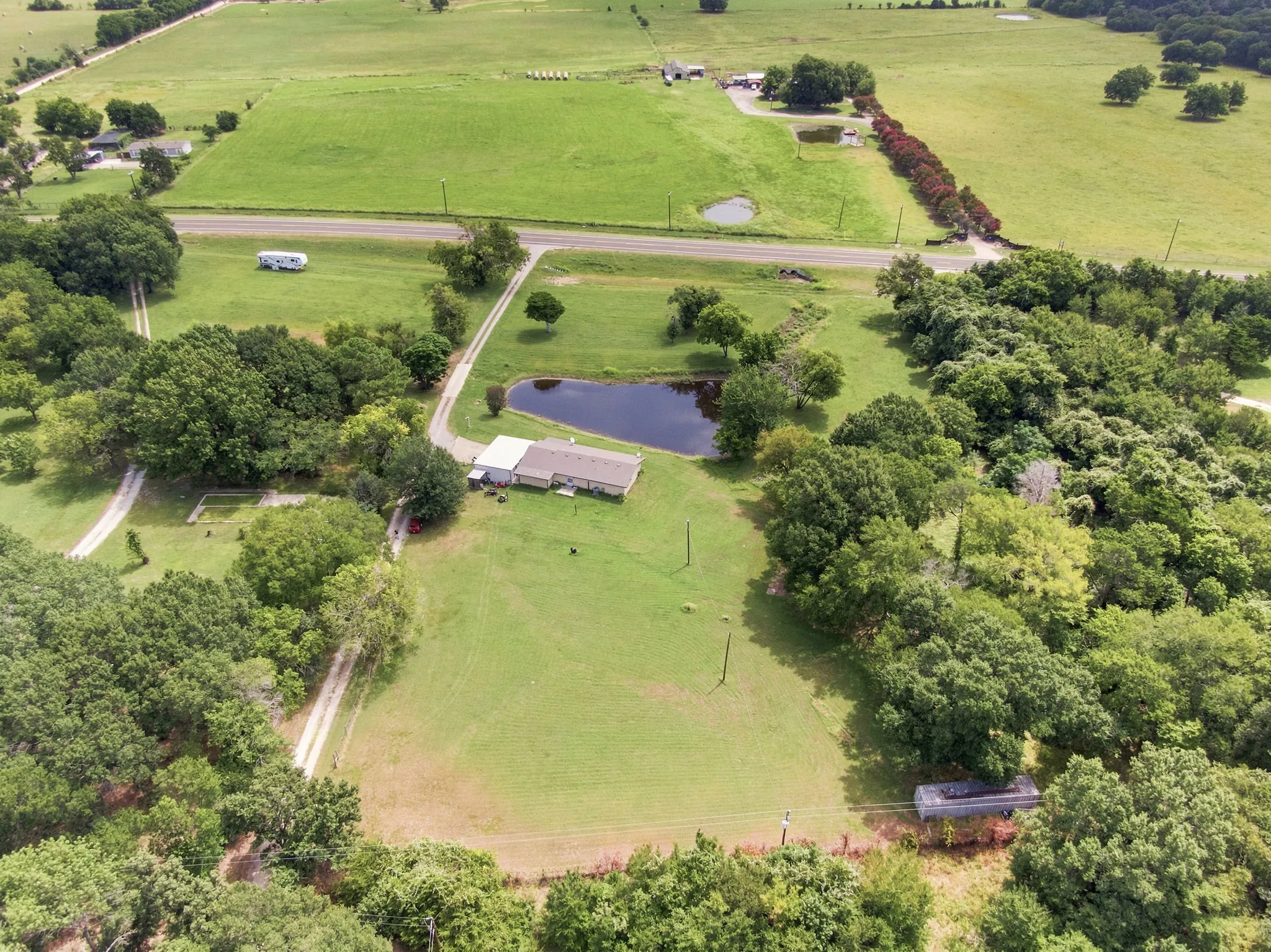 Expansive property featuring a single-story residence with a light-colored roof, multiple ponds, a paved driveway, mature trees, and open green fields