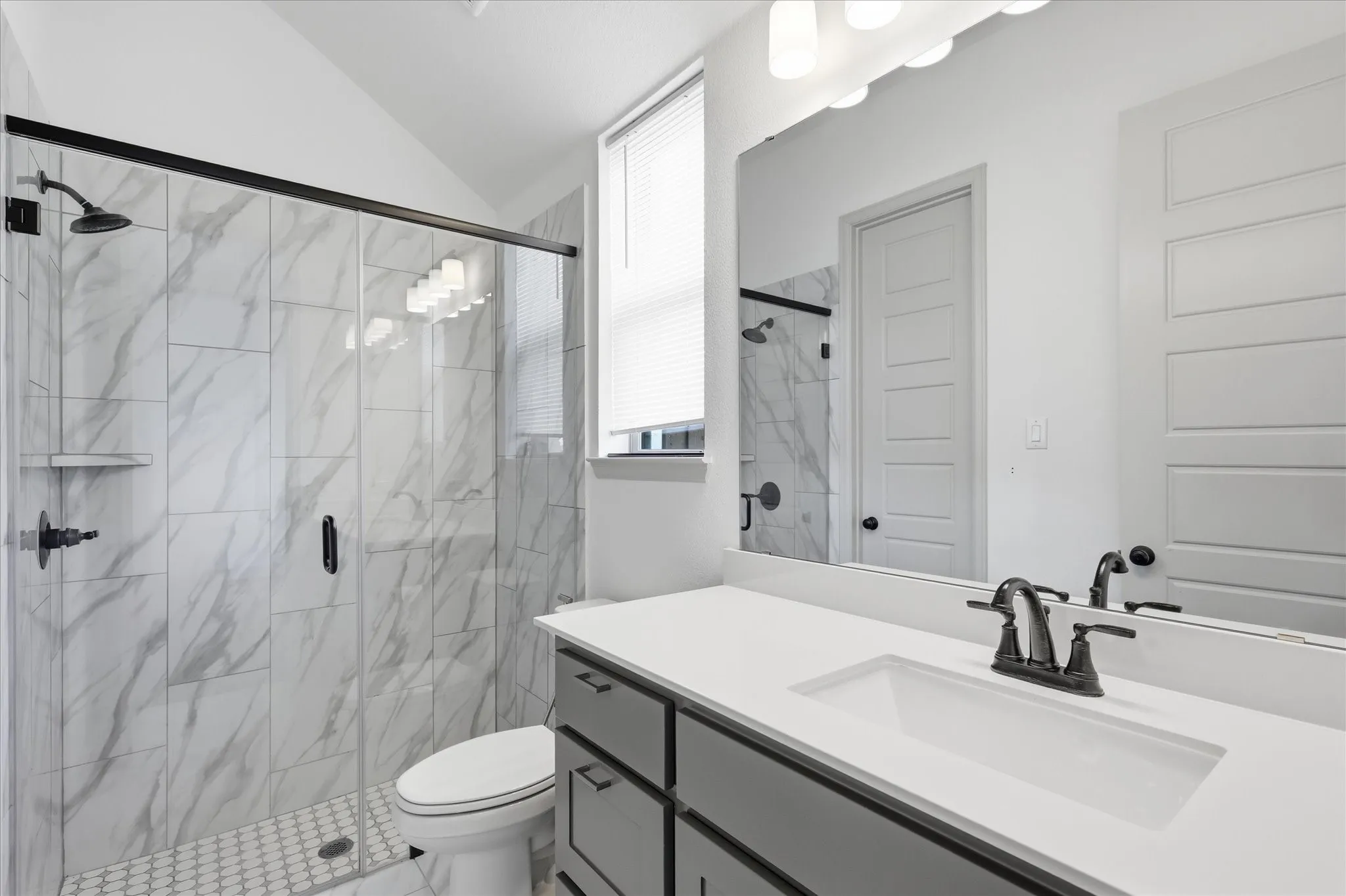 Bathroom featuring a walk-in shower with large format tile surround and glass enclosure, a white vanity with integrated sink, chrome-finish fixtures, and built-in vanity lighting