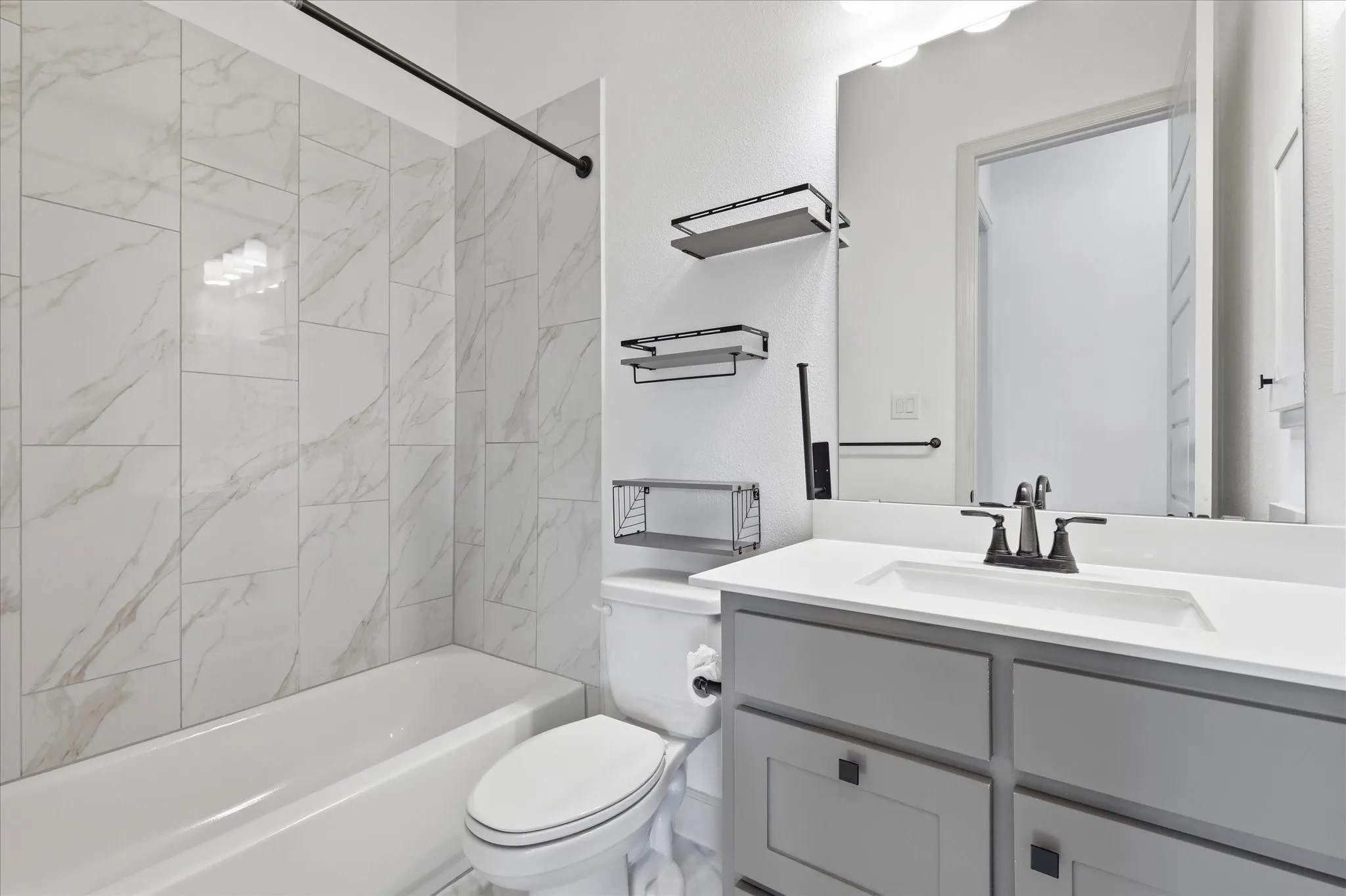 Bathroom featuring a white bathtub, large format tile surround with marble-look veining, gray vanity with dark hardware, white countertop with integrated sink, and dark-finished faucet