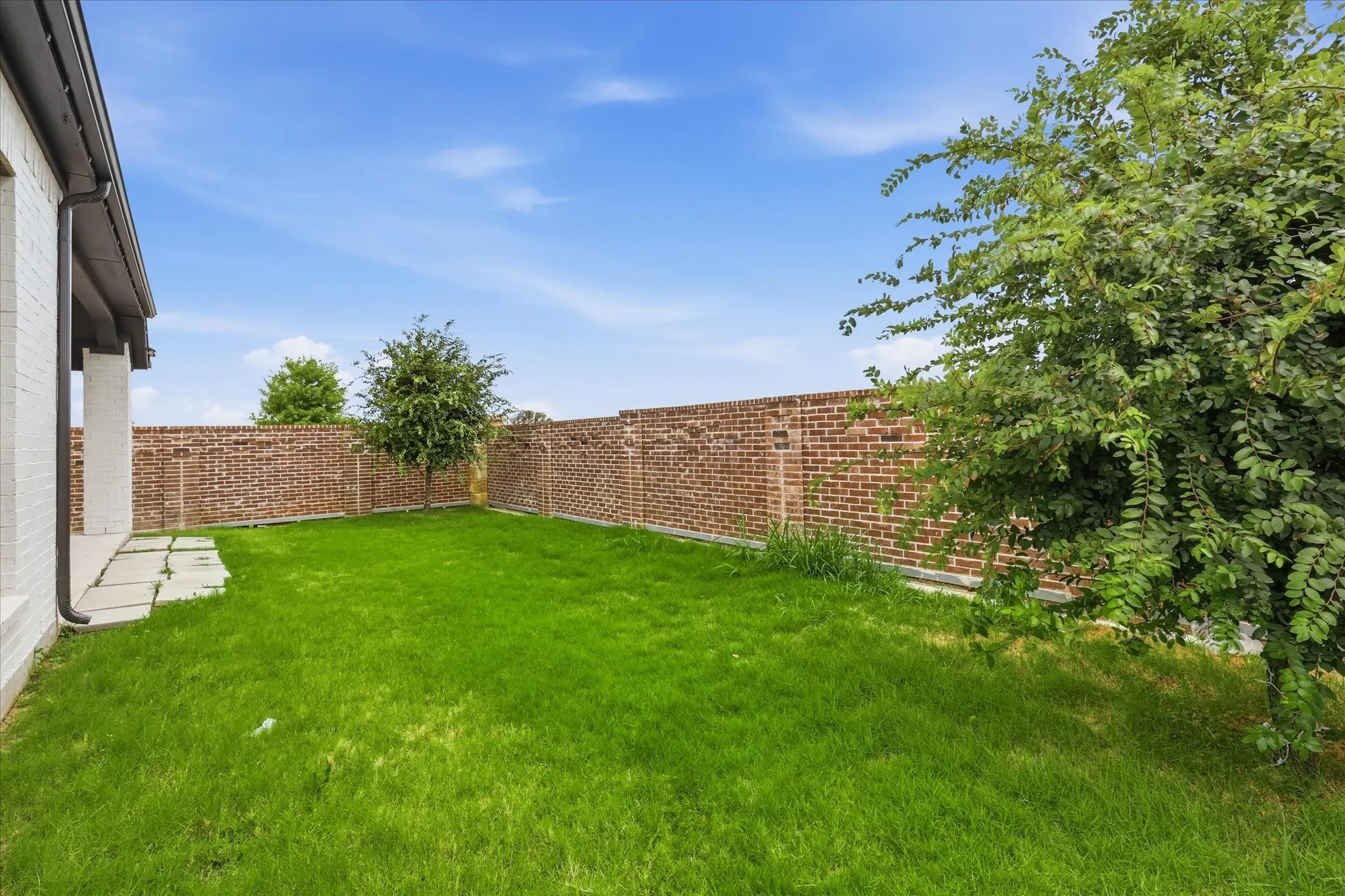 Spacious grassy yard featuring a brick perimeter wall, mature trees, and a flagstone patio area