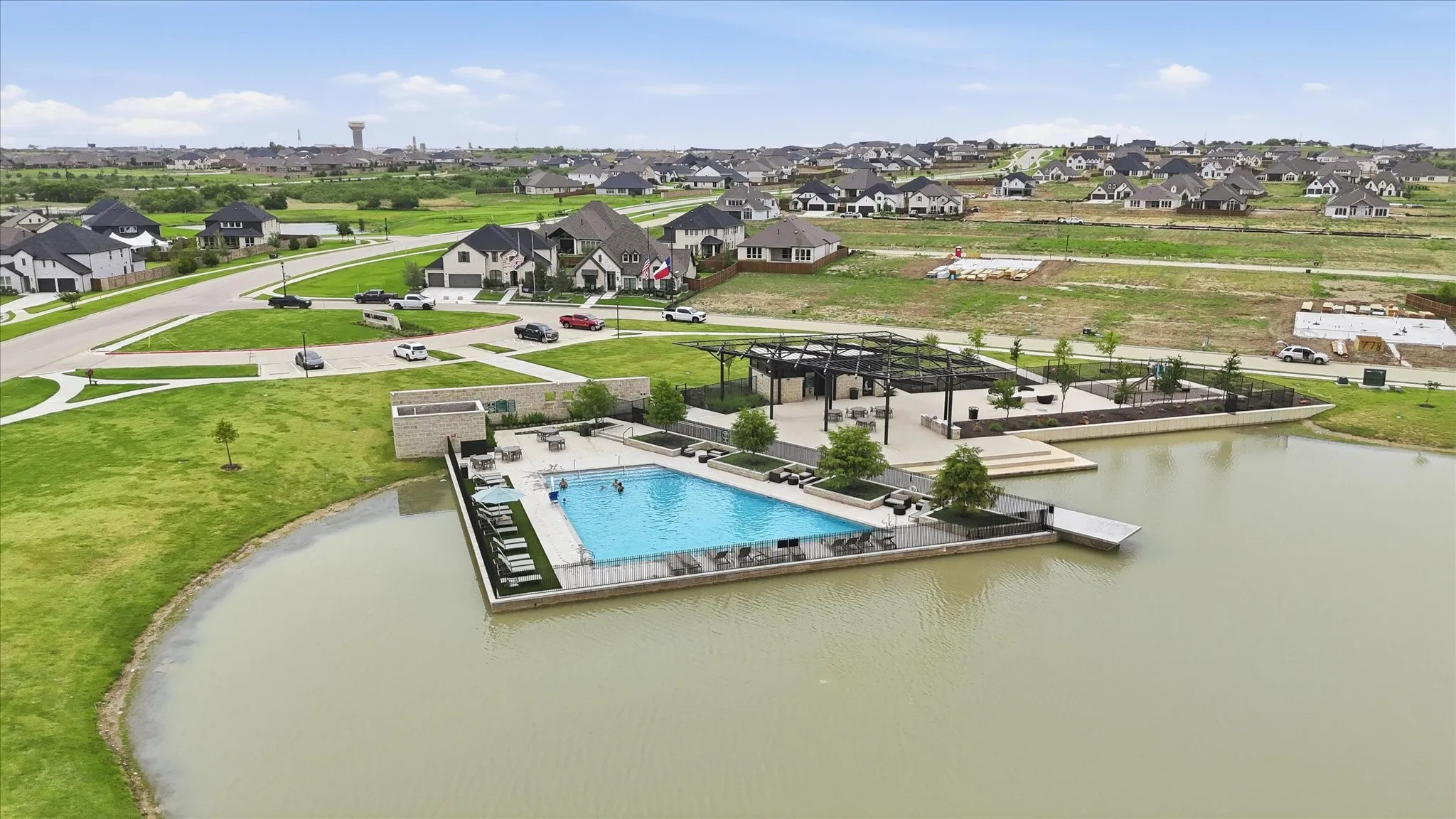 Community pool area featuring a rectangular swimming pool with surrounding lounge seating, a dock extending into the lake, and a covered patio structure with pergolas