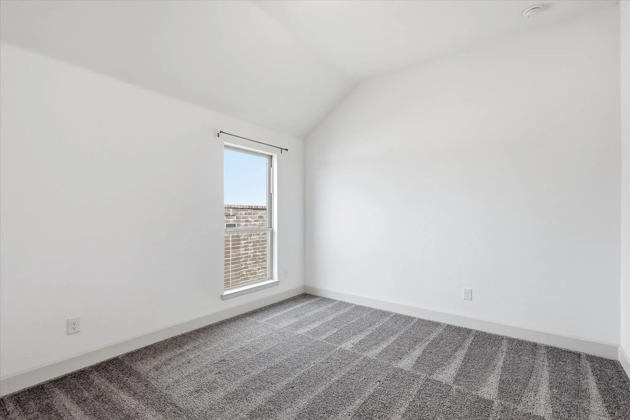 Room featuring a window with a brick exterior visible, clean white walls, a textured gray carpet, and a sloping ceiling line