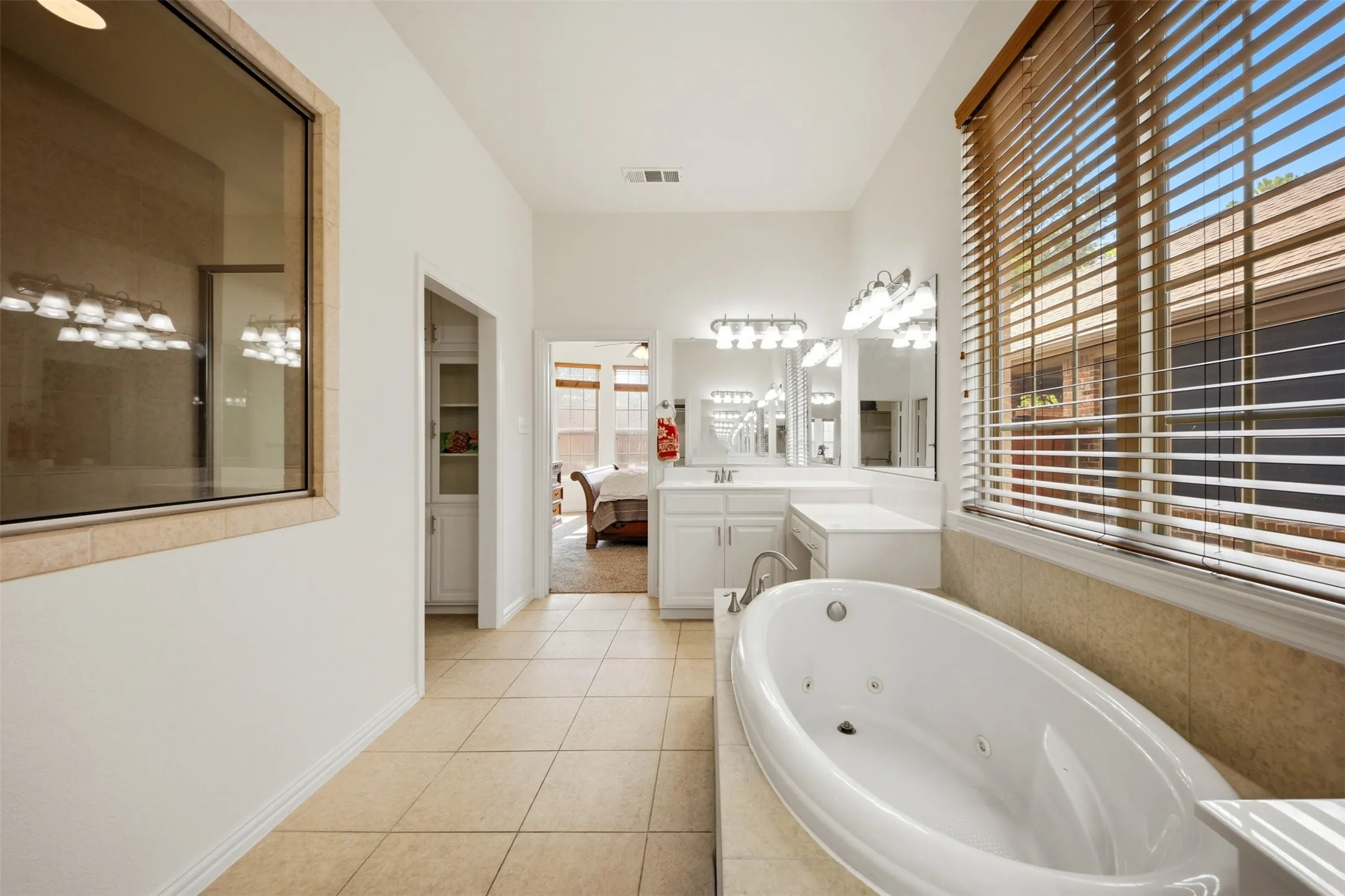 White jetted soaking tub, dual vanity with individual mirrors, wall-mounted lighting fixtures, natural light from a large window, and light-toned tile flooring