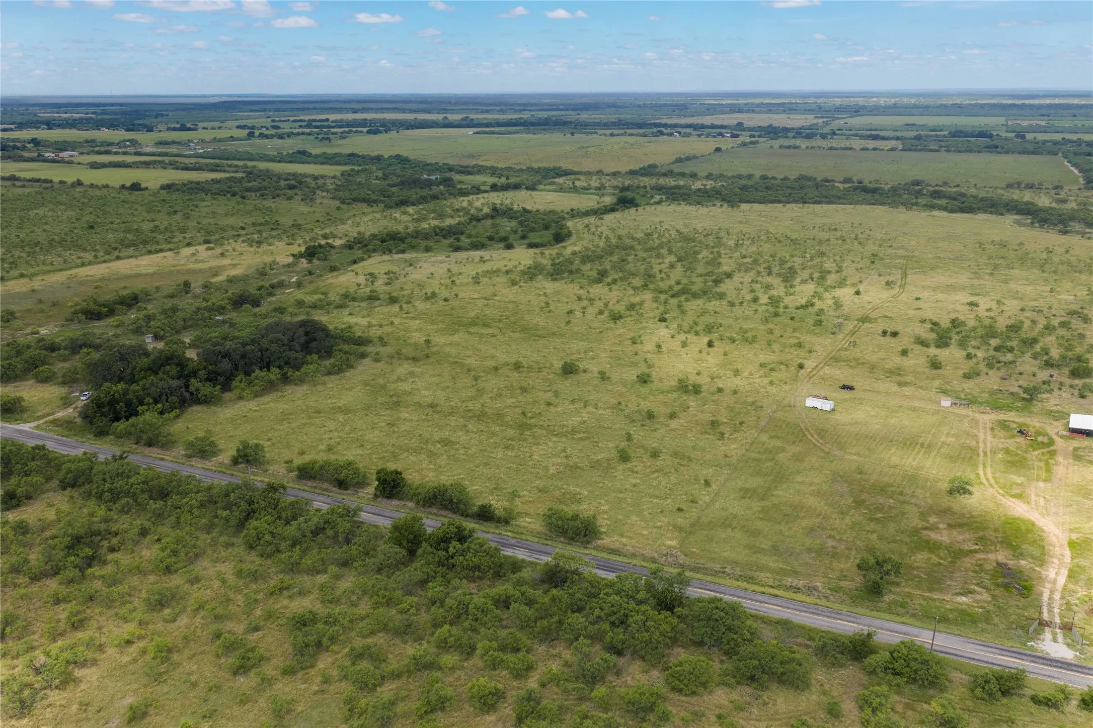 Expansive rural acreage featuring a paved road frontage, natural vegetation, and distant agricultural structures