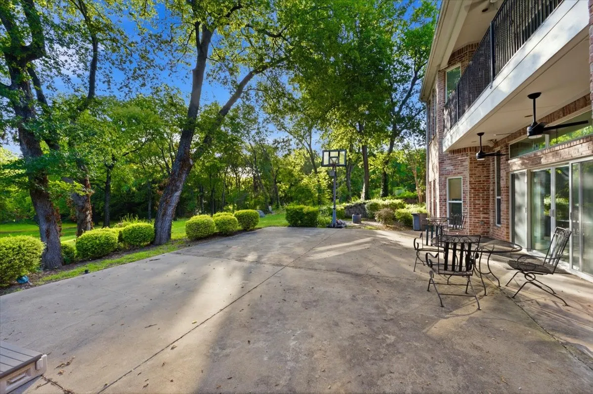 Expansive concrete patio bordered by mature trees and manicured shrubs