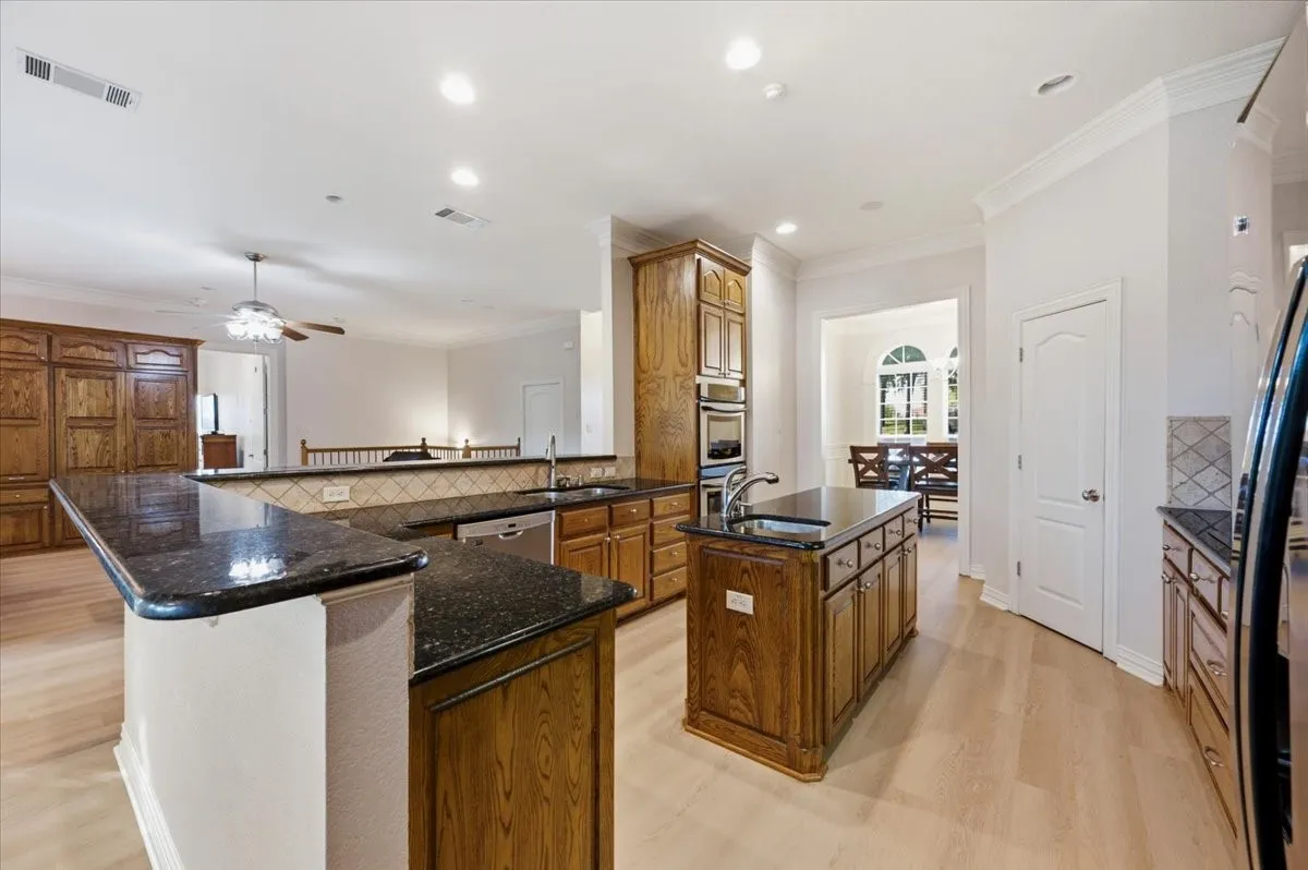 Kitchen featuring wood-finish cabinetry, dark stone countertops, a central island with an integrated sink, built-in stainless steel appliances, and light wood-finish flooring