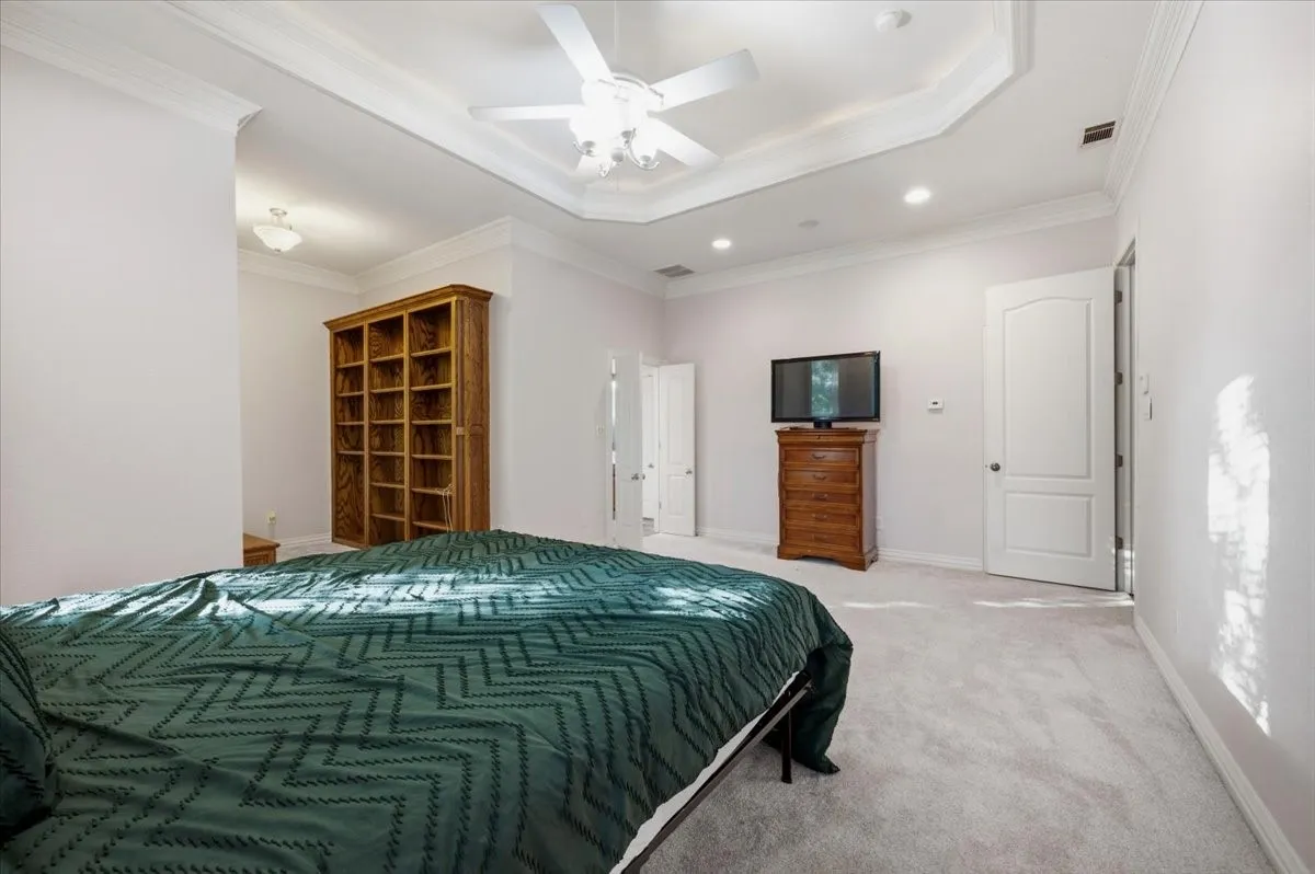Spacious room featuring light-colored carpeting, a tray ceiling with crown molding and a ceiling fan, recessed lighting, and a built-in bookshelf