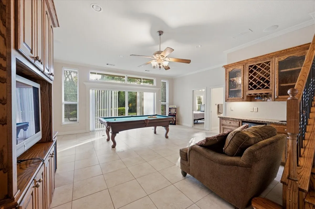 Spacious room featuring light-toned tile flooring, a built-in wet bar with wood cabinetry and countertop, a ceiling fan, and large windows with vertical blinds