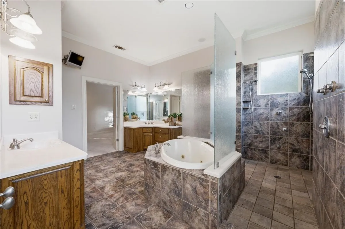 Spacious bathroom featuring a jetted tub, a separate tiled shower with a glass partition, dual vanity sinks with wood-finish cabinetry, and tiled flooring