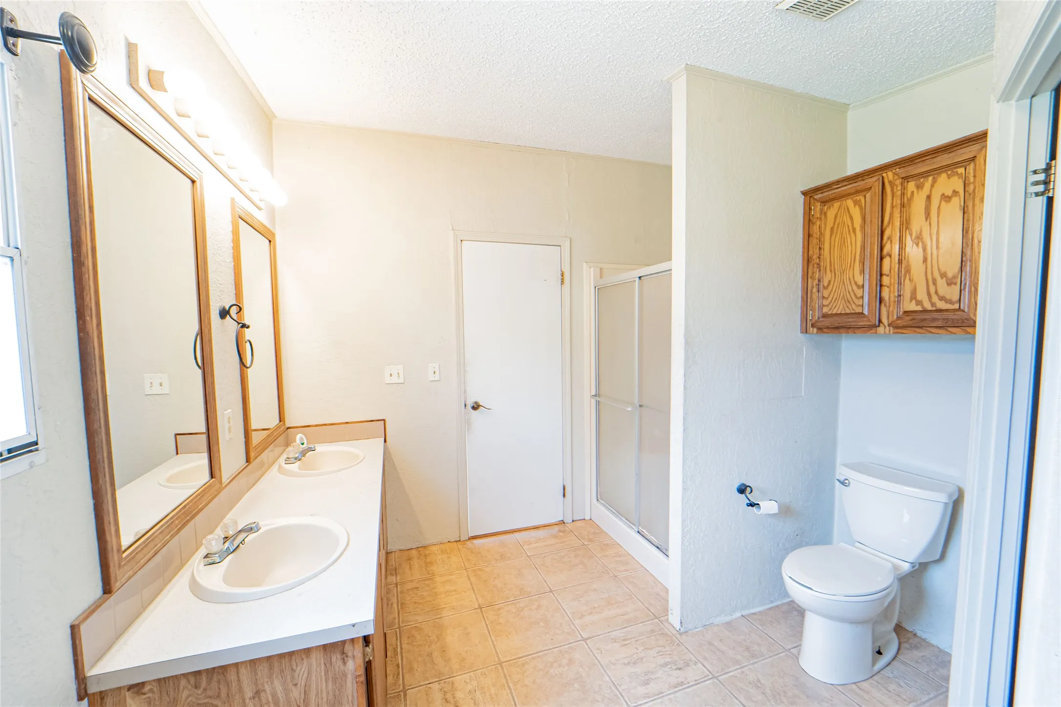 Spacious bathroom featuring a double vanity with wood-finish cabinetry, a tiled shower with a sliding glass door, a toilet, and overhead wooden cabinetry