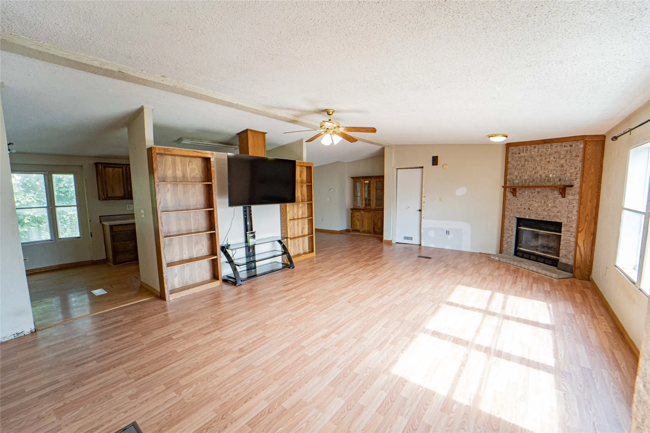 Spacious living area featuring wood-finish flooring, a stone-faced fireplace with a wood mantel, built-in shelving, a ceiling fan, and multiple windows for natural light