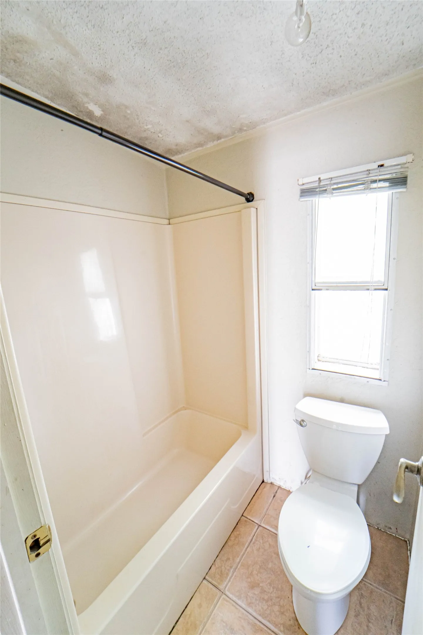 Bathroom featuring a white tub-shower combination, a white toilet, and a single-pane window with blinds