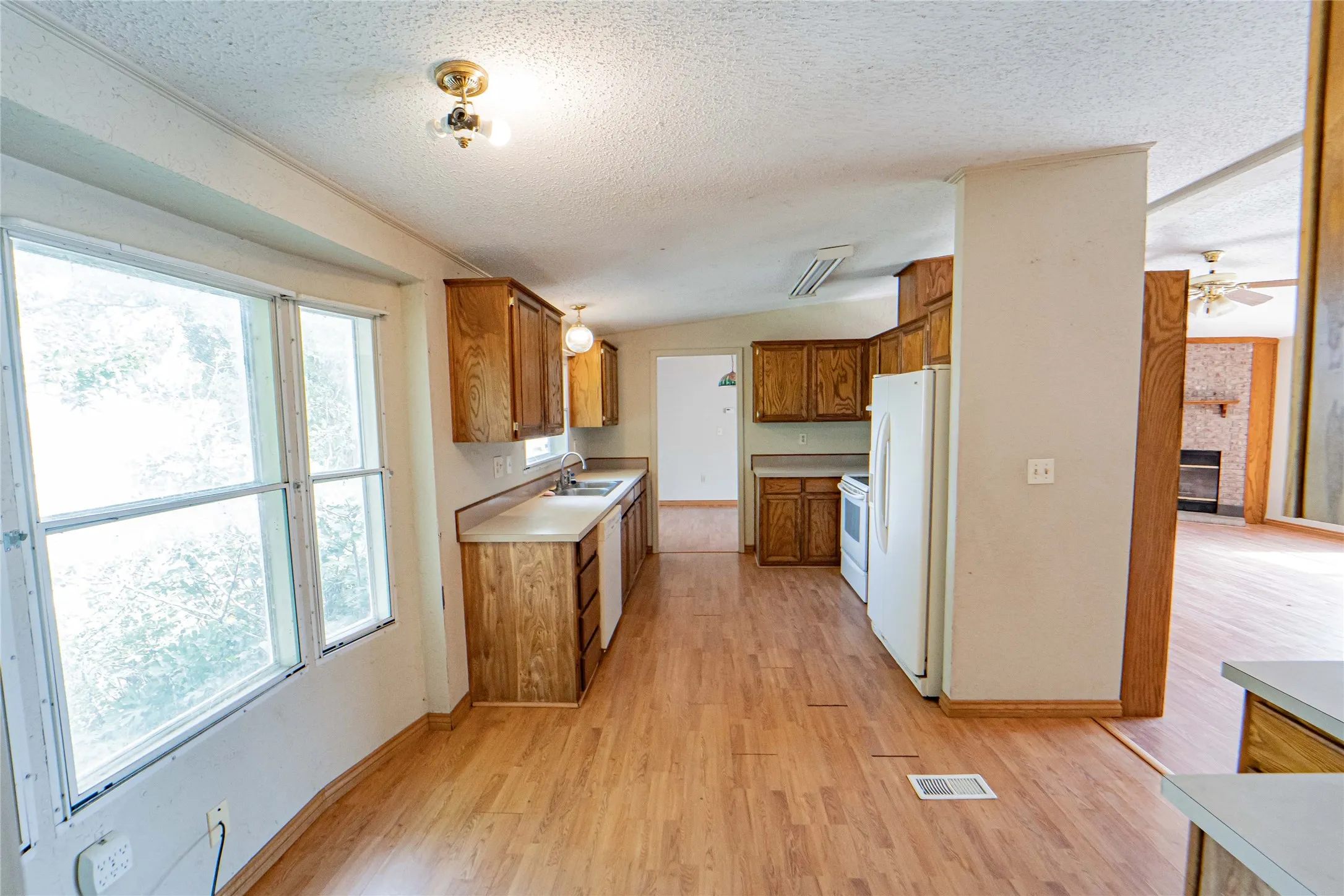 Kitchen , open to dining, featuring wood-finish flooring, wood cabinetry, light-toned countertops, a stainless steel sink, and white appliances