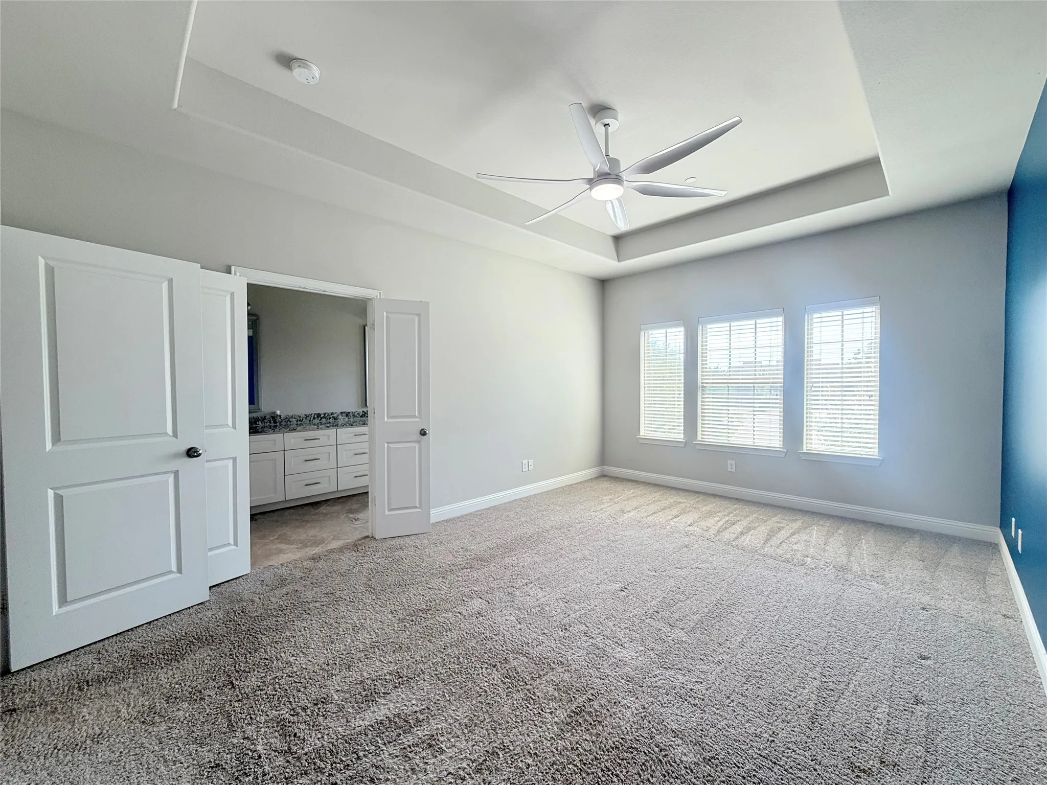 Carpeted room featuring a tray ceiling with a modern ceiling fan, three windows with blinds, and white double doors opening to a bathroom with a dual vanity and granite-finish countertop