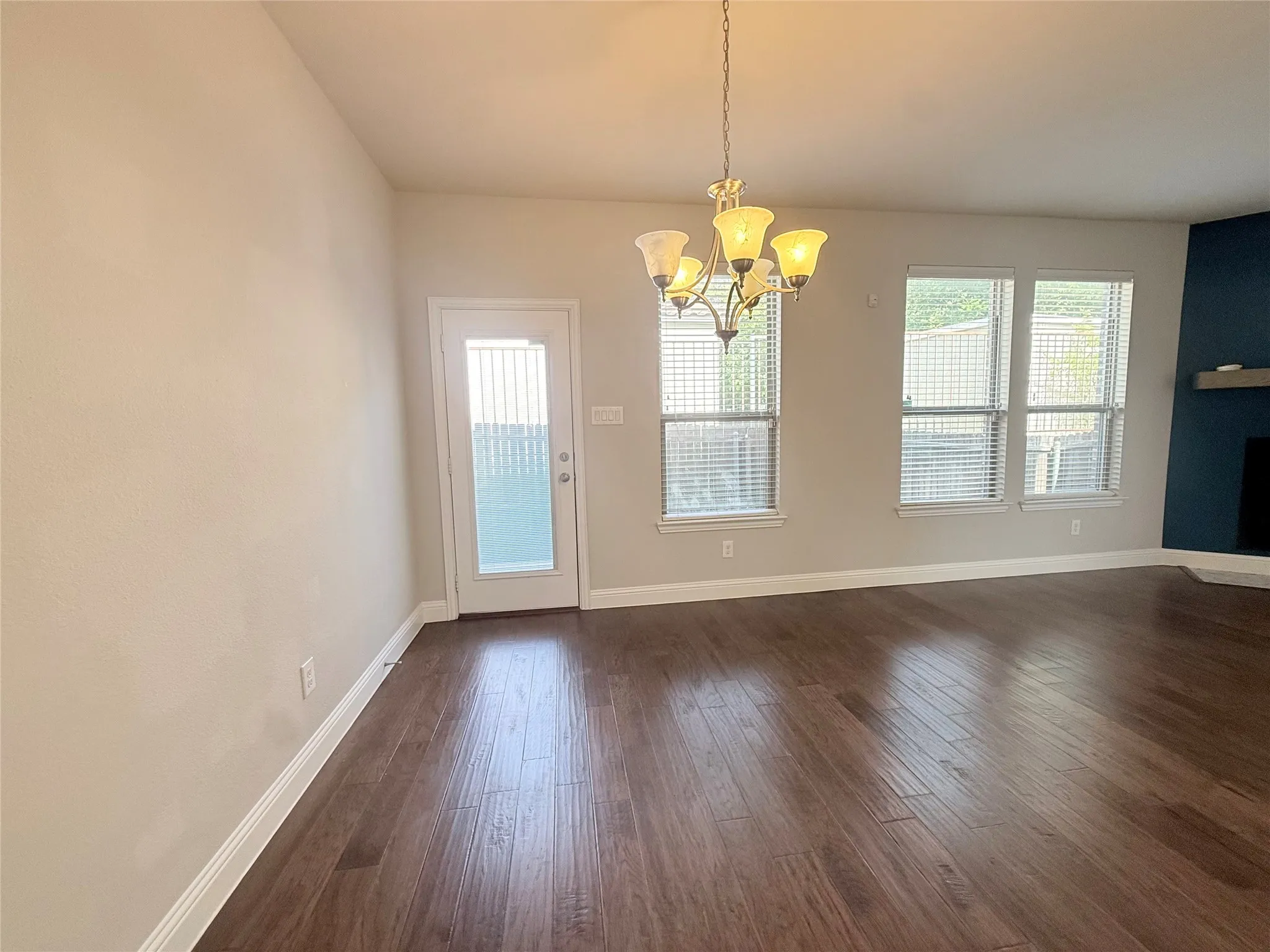 Spacious room featuring wood-finish flooring, a contemporary chandelier, multiple windows with blinds, a glass-paned exterior door, and a corner fireplace with a mantel