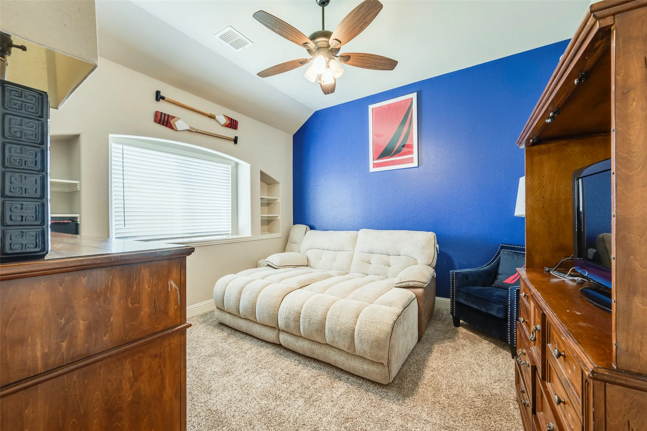 Sloped ceiling featuring a ceiling fan, recessed wall shelving, and a prominent window with blinds