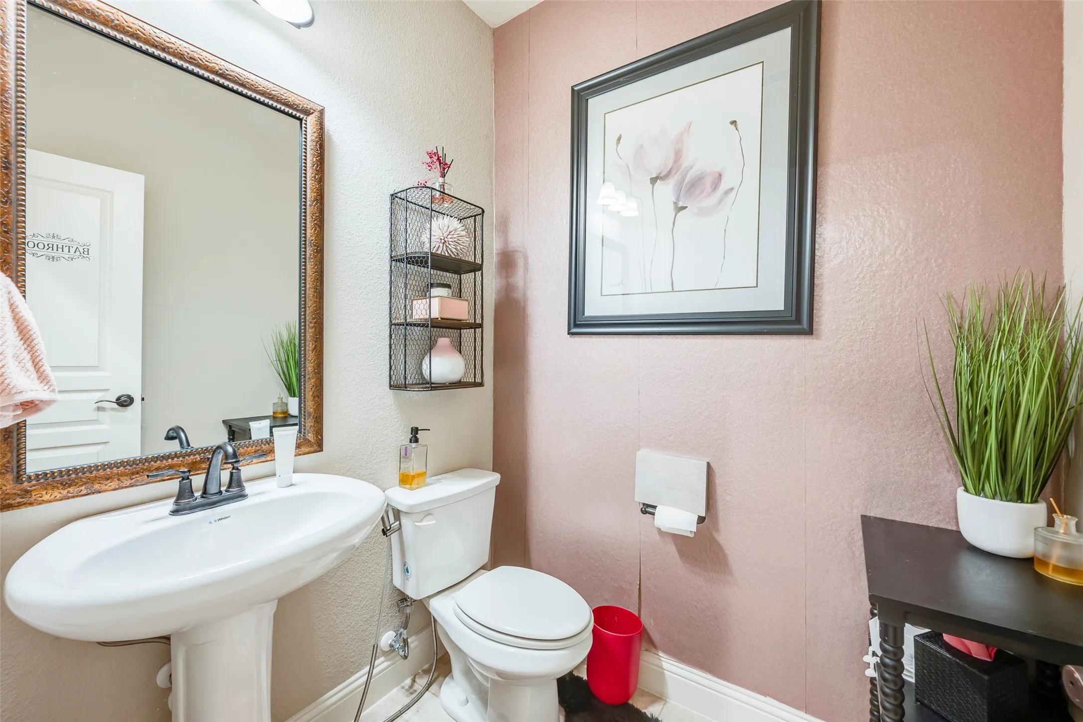 Pedestal sink with a dark bronze-finish faucet, framed mirror with ornate detailing, and a white toilet