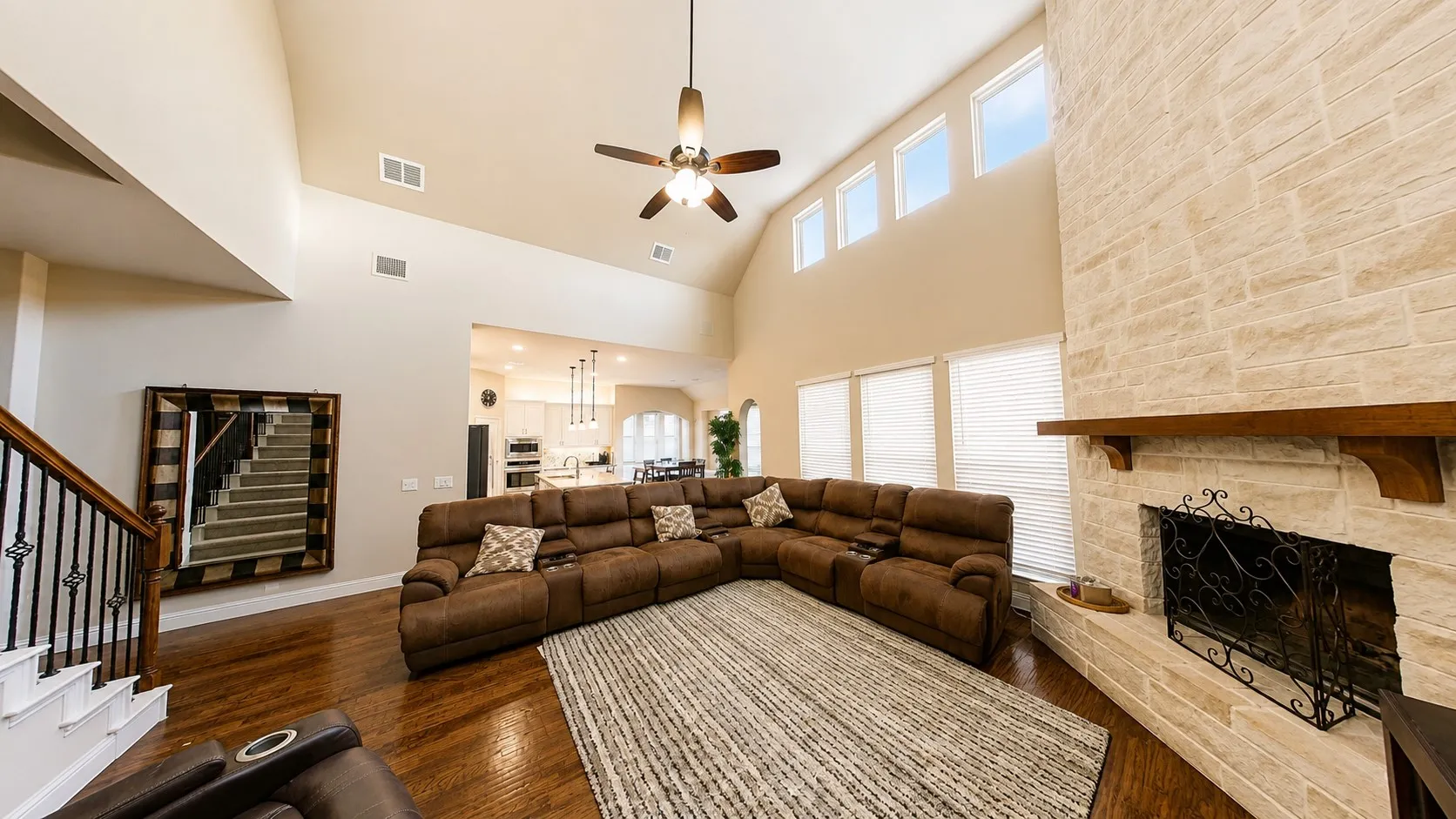 Vaulted ceiling with a ceiling fan, stone fireplace with a wood mantle, wood-finish flooring, and a staircase with wrought iron balusters