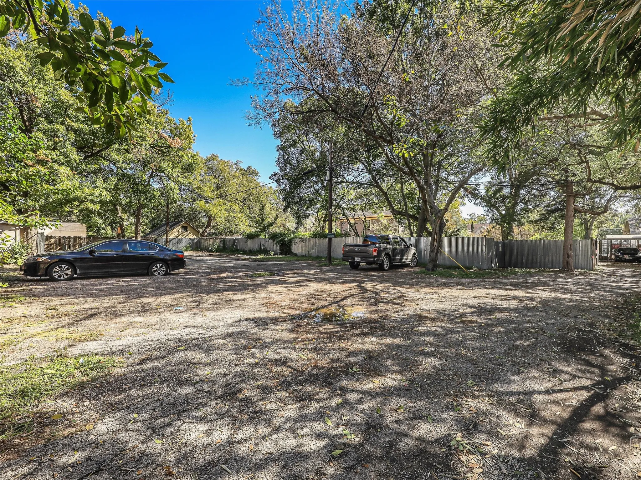 Expansive gravel parking lot surrounded by mature trees, featuring a wood fence, and partial tree canopy coverage