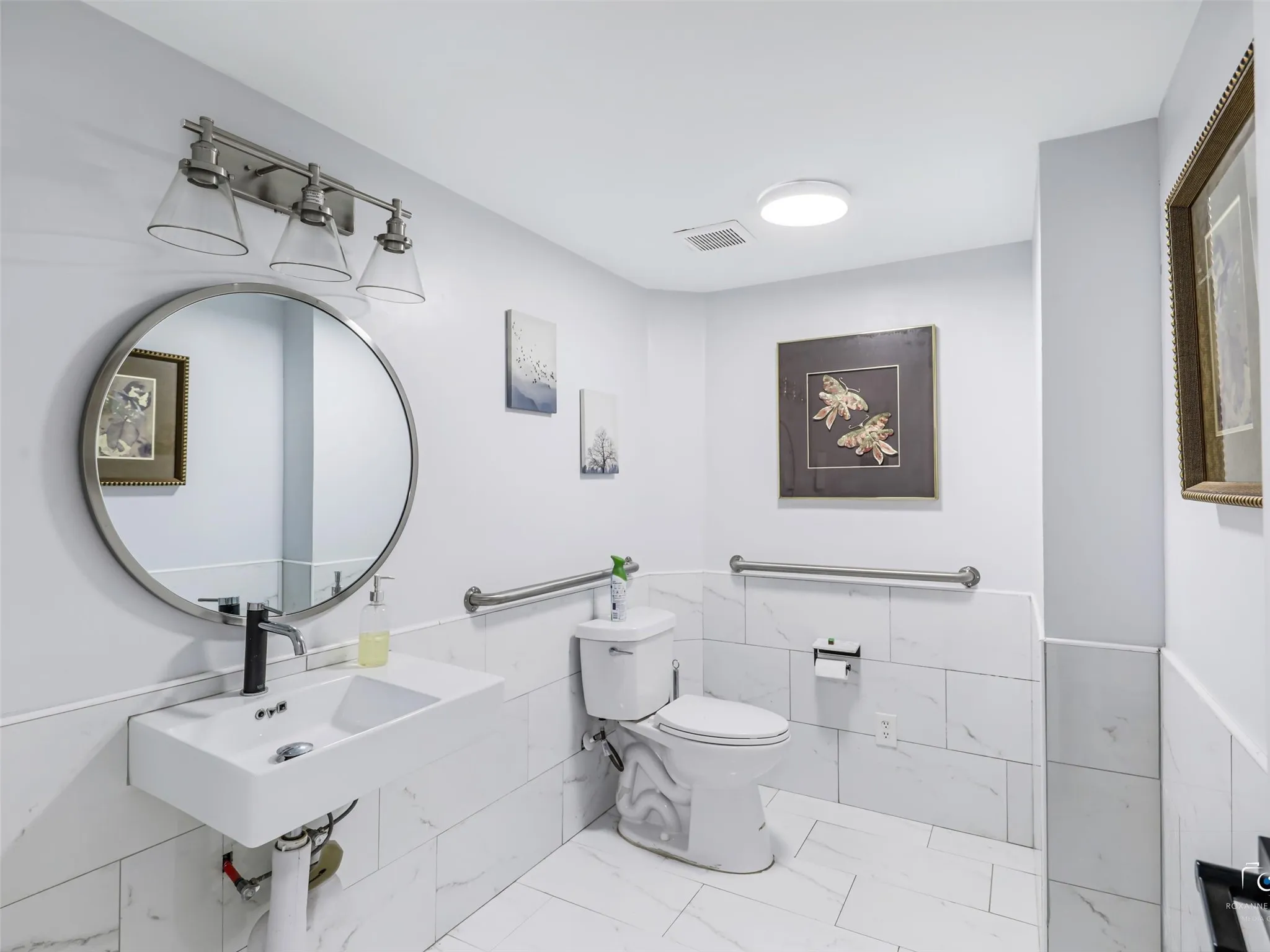 Bathroom featuring white tile flooring and wainscoting, a wall-mounted rectangular sink, a circular mirror with a brushed nickel frame, a three-light vanity fixture, and a toilet with grab bars