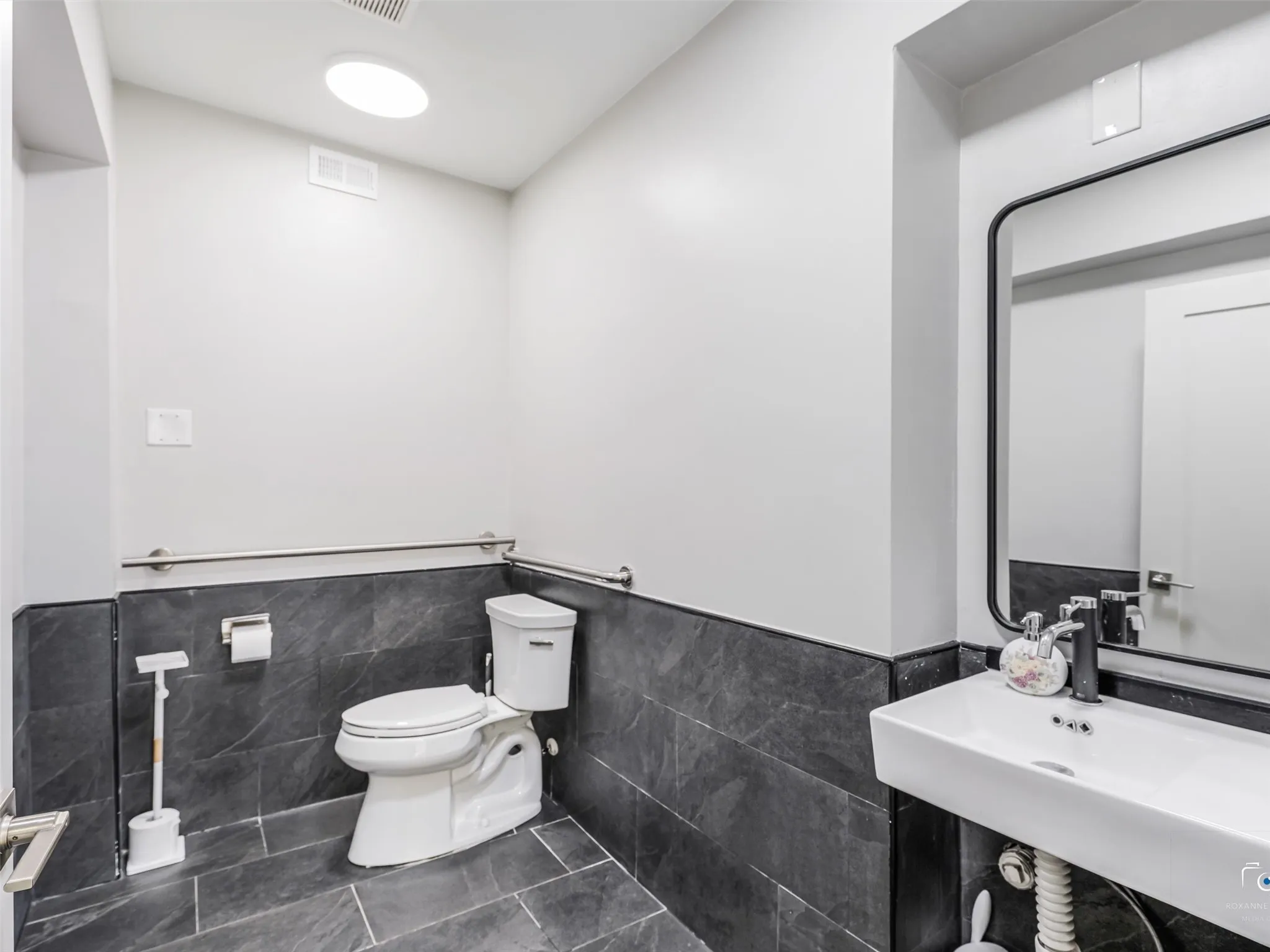 Modern bathroom featuring dark-toned tile flooring and wainscoting, a wall-mounted rectangular sink, and a contemporary black-framed mirror