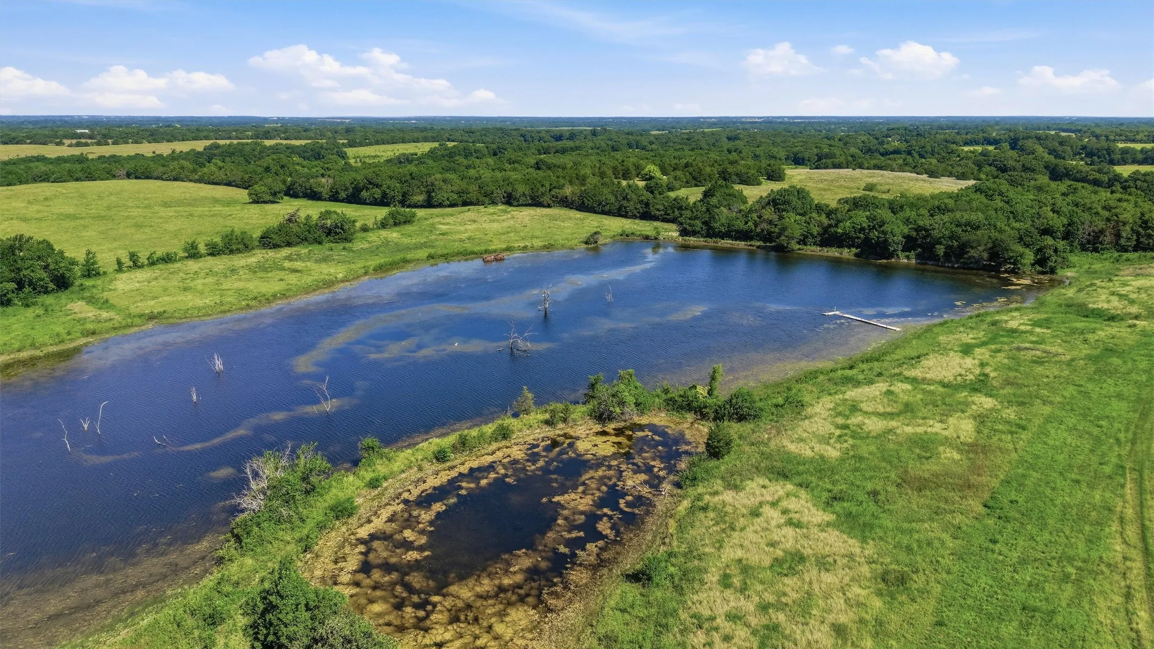 Expansive acreage featuring a large pond with a dock, surrounded by lush green fields and mature tree lines