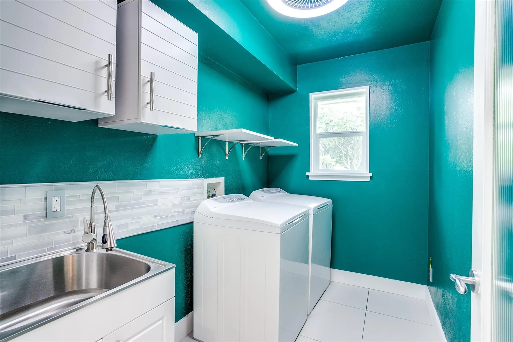 Dedicated utility space featuring a stainless steel sink, white shaker-style cabinetry, and white horizontal tile backsplash