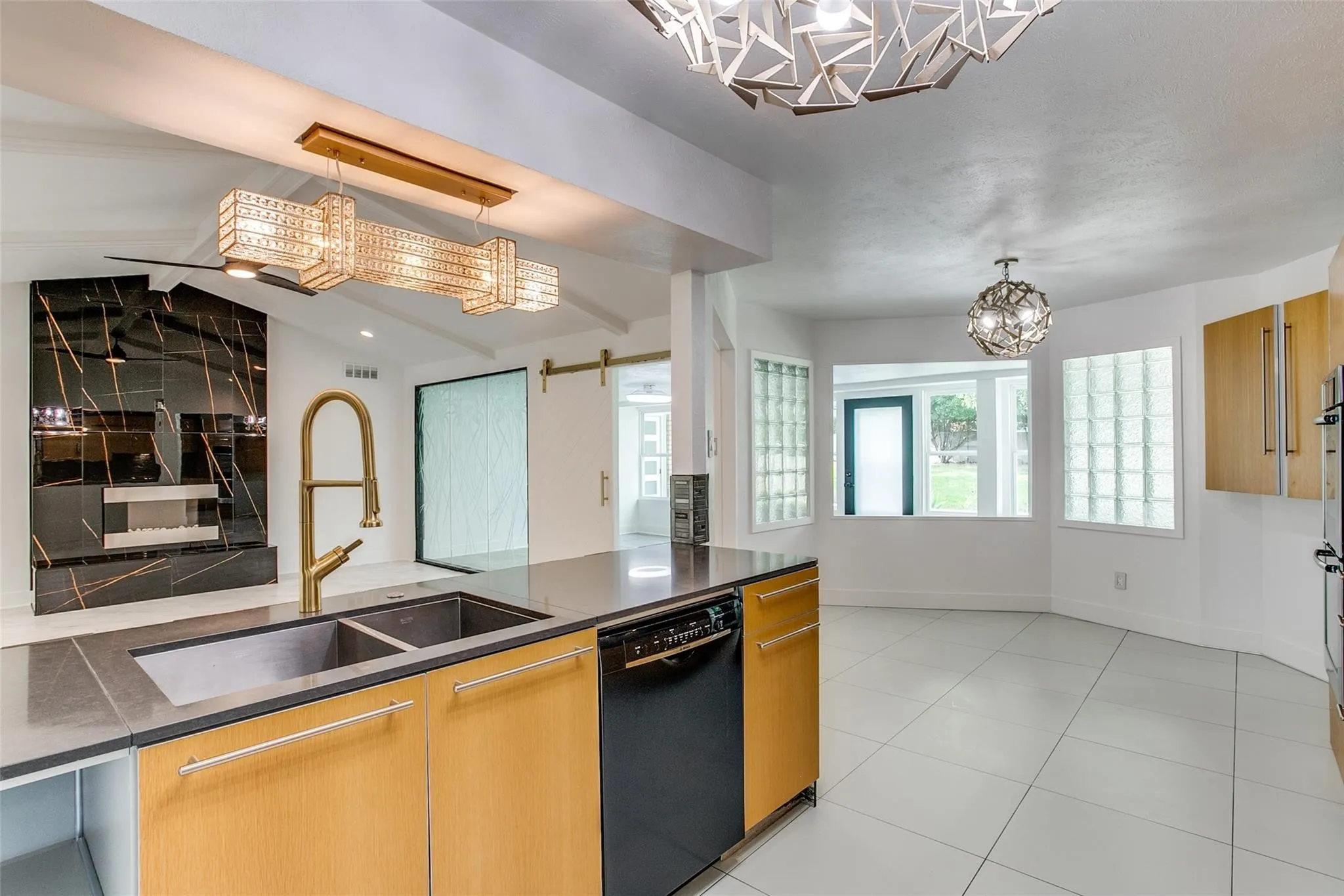 Contemporary kitchen featuring an island with a dual-basin sink and brushed gold faucet, light wood-finish cabinetry, built-in dishwasher, and a dark countertop