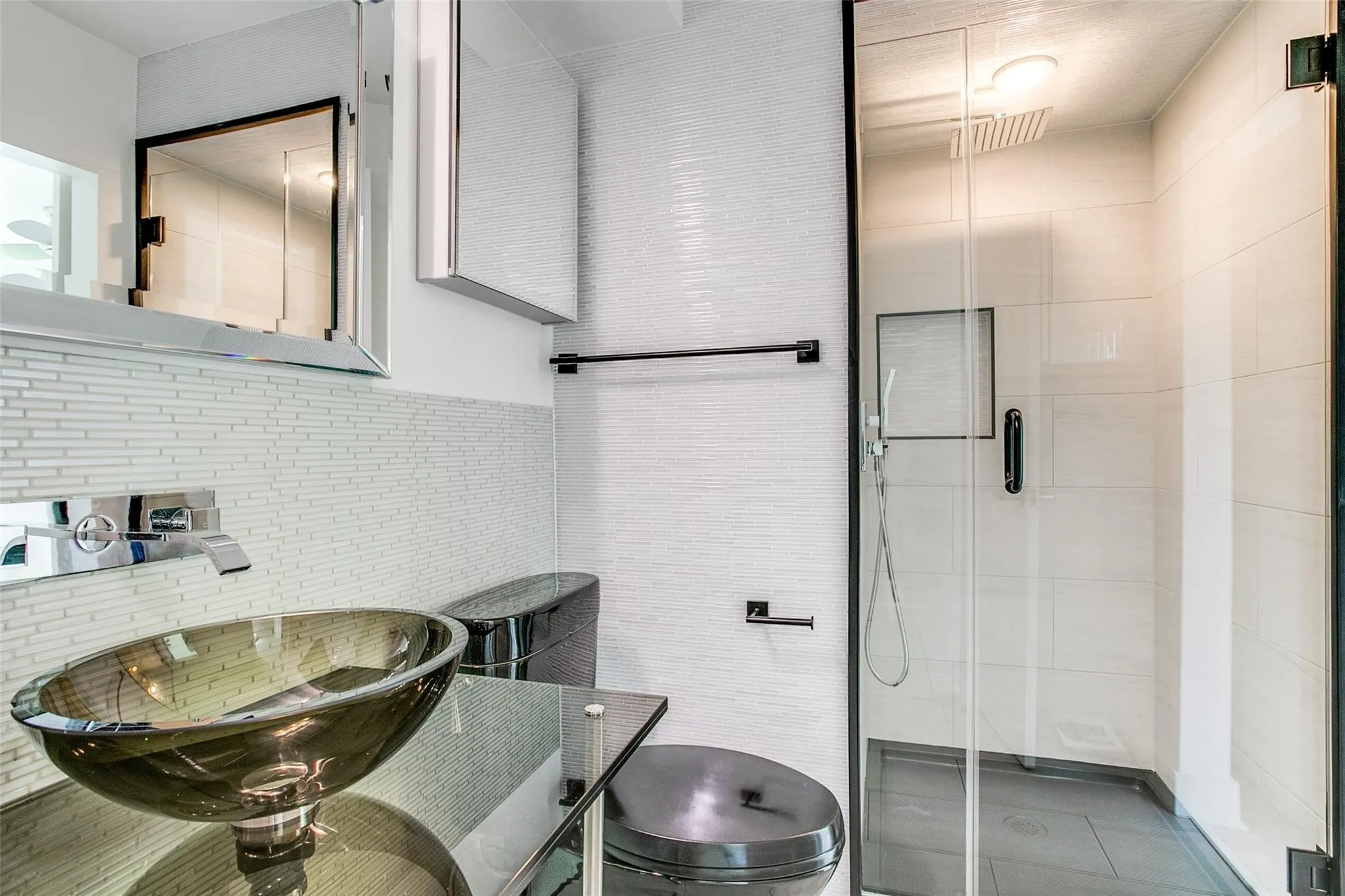 Contemporary bathroom featuring a vessel sink on a glass countertop, a wall-mounted faucet, a black toilet, a glass-enclosed shower with a rainfall showerhead, and textured white wall tile