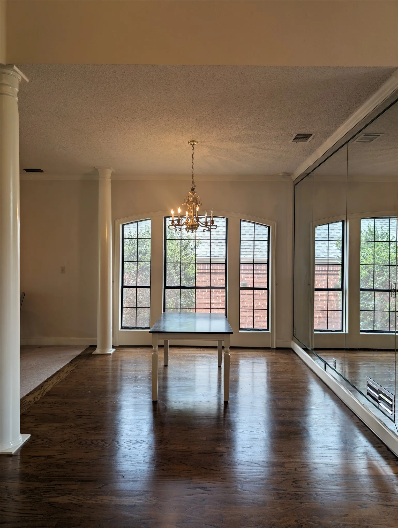 Spacious formal dining room featuring dark wood-finish flooring, architectural columns, a brass chandelier, and arched windows with horizontal blinds