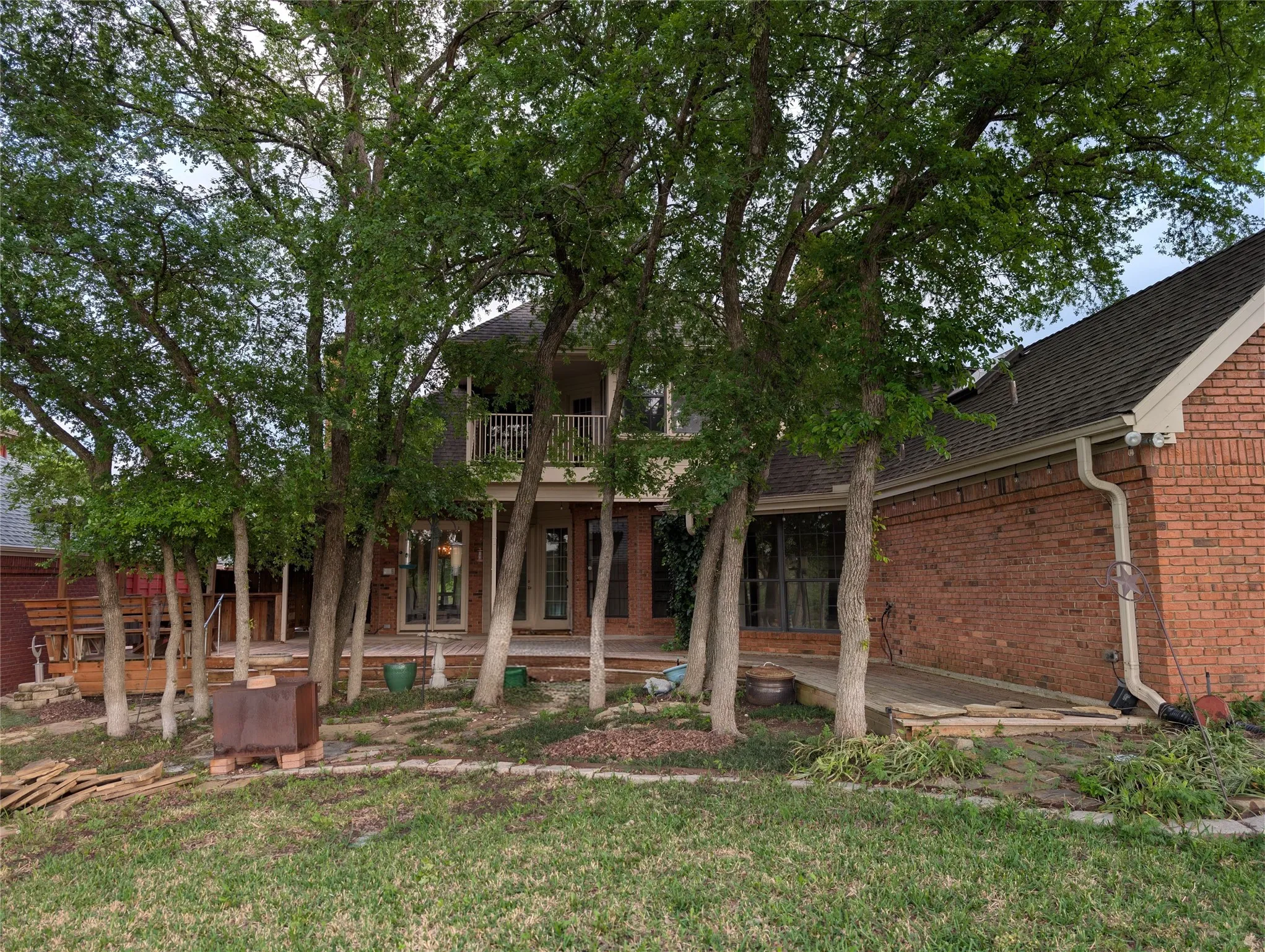 Brick exterior residence featuring mature trees, a second-story balcony with metal railing, and a ground-level patio with multiple French doors