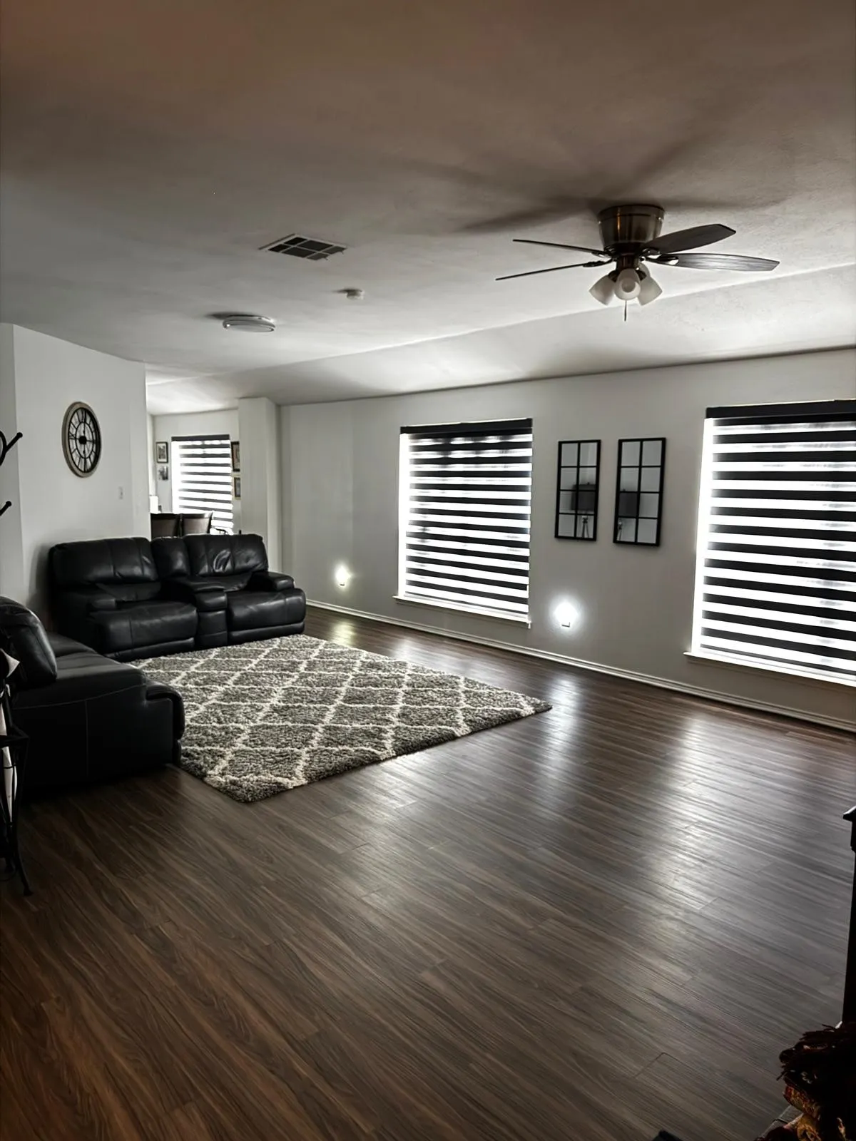 Spacious living area featuring dark wood-finish flooring, multiple windows with blinds, and a ceiling fan with integrated lighting