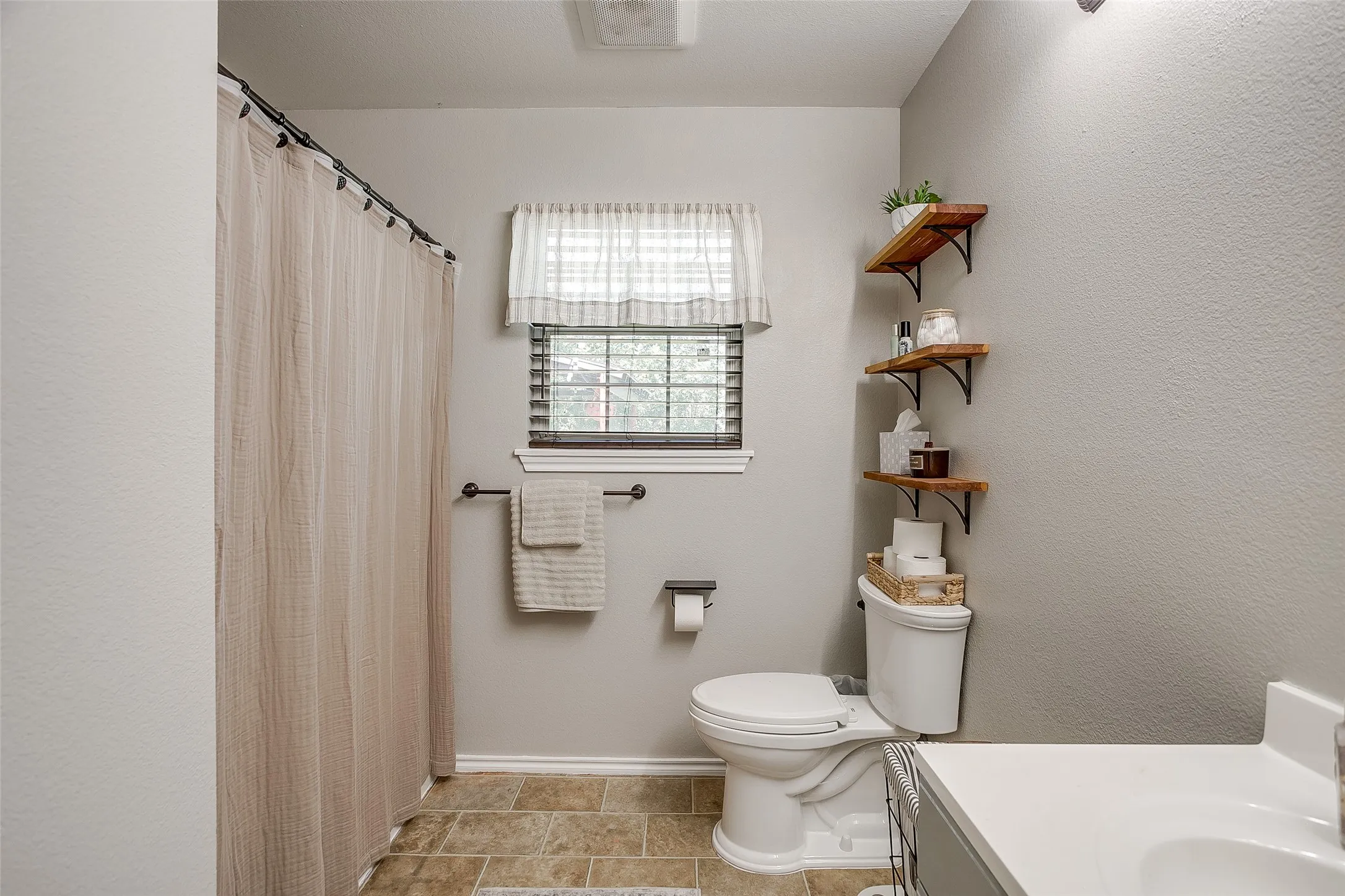 Bathroom featuring a window with blinds, tile flooring, a toilet, and wall-mounted shelving