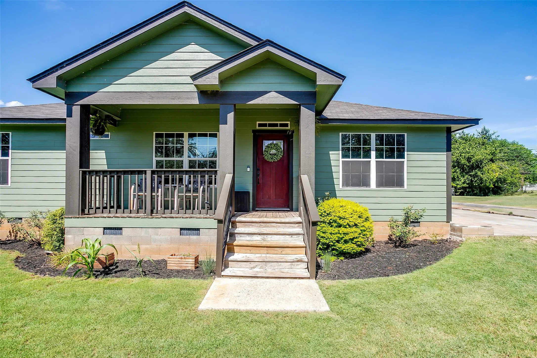 Green siding exterior with a dark red entry door, a covered porch with dark wood railings, and a concrete walkway