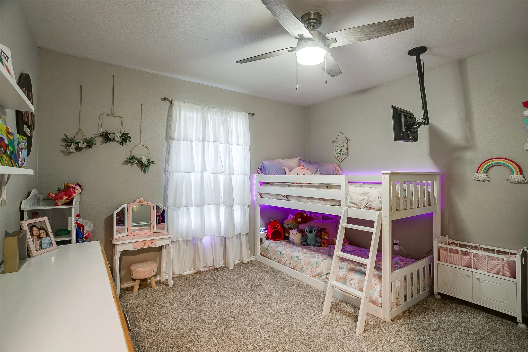 Carpeted interior room featuring a ceiling fan with light fixture, built-in shelving, mounted television bracket, a window with sheer curtains, and light gray wall paint