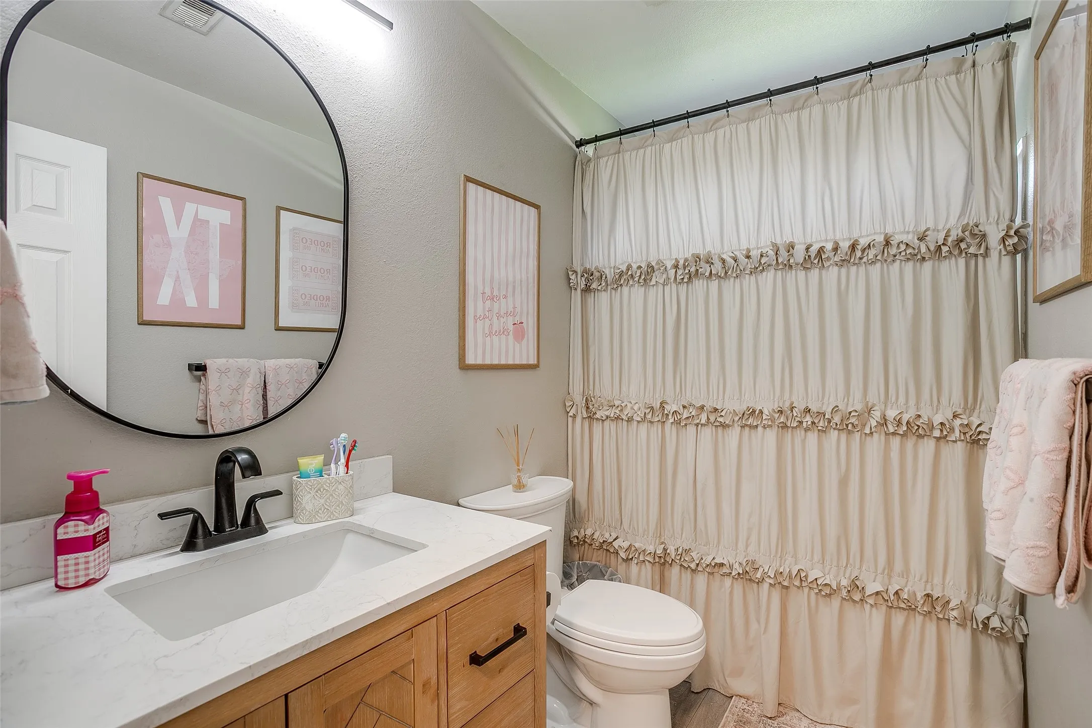 Bathroom featuring a wood-finish vanity with a white countertop and integrated sink, a matte black faucet, and a round black-framed mirror