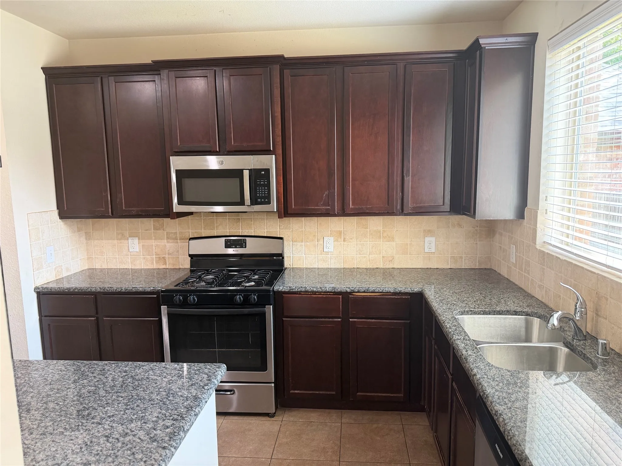 Kitchen featuring dark wood cabinetry, speckled countertops, tile backsplash, and stainless steel appliances including a gas range, microwave, and double basin sink