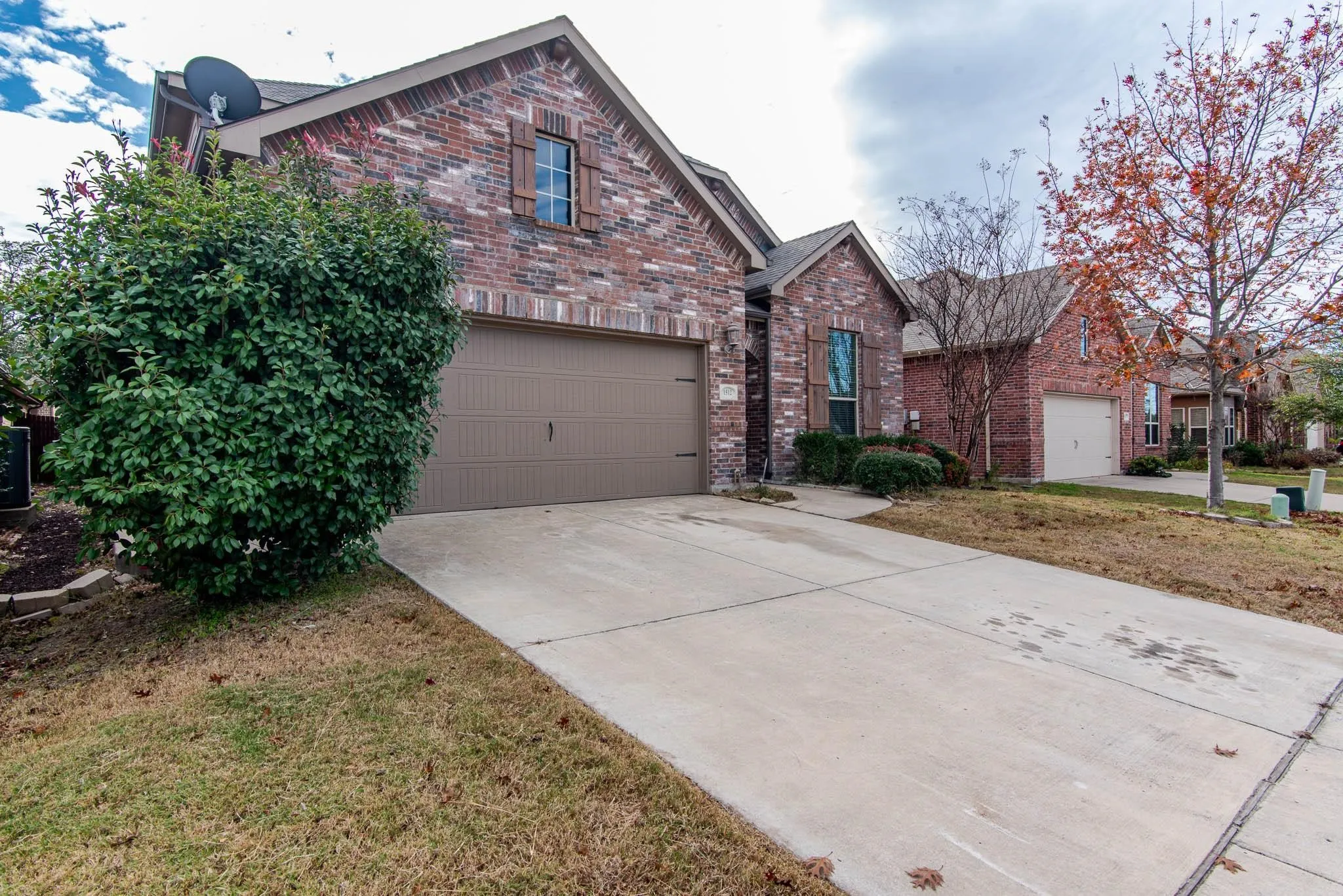 Brick facade home featuring an attached garage with a tan door, a concrete driveway, and mature landscaping