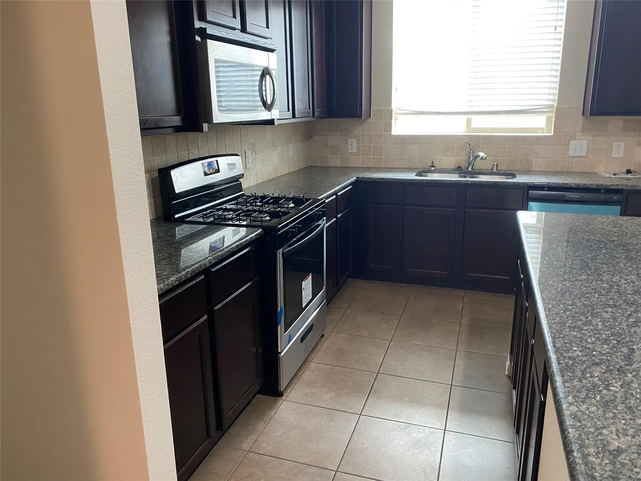Kitchen featuring dark wood cabinetry, a gas range with stainless steel accents, a built-in microwave, granite countertops, and a tiled backsplash