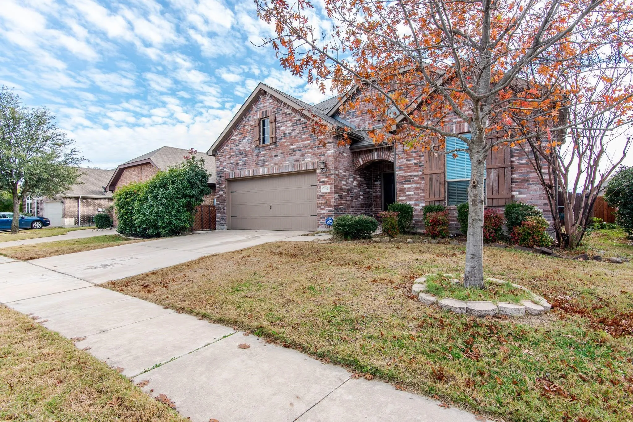 Brick facade residence featuring an attached two-car garage, mature front yard trees, decorative window shutters, and a concrete driveway