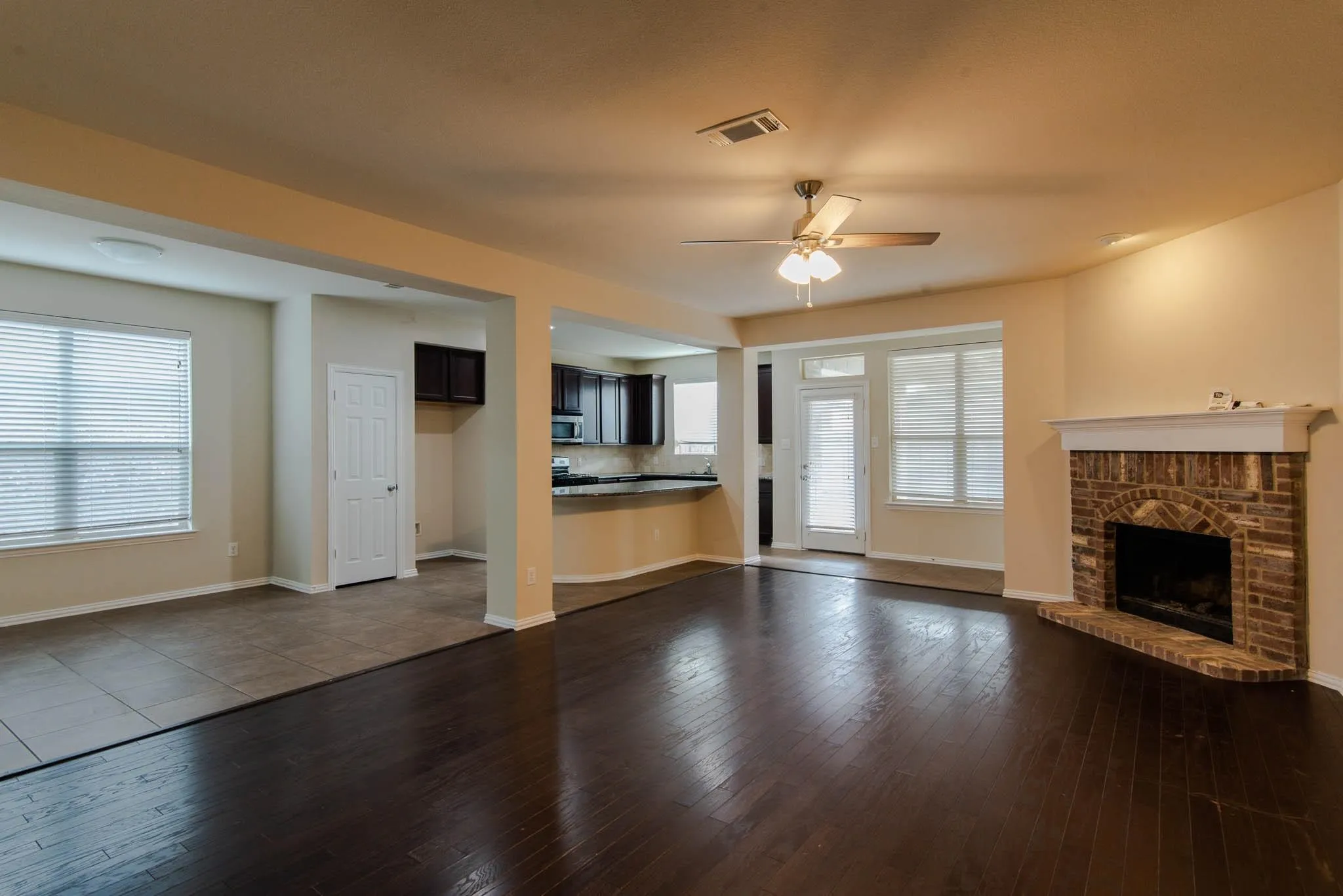 Spacious living area featuring dark wood-finish flooring, a brick fireplace with a white mantel, and a ceiling fan