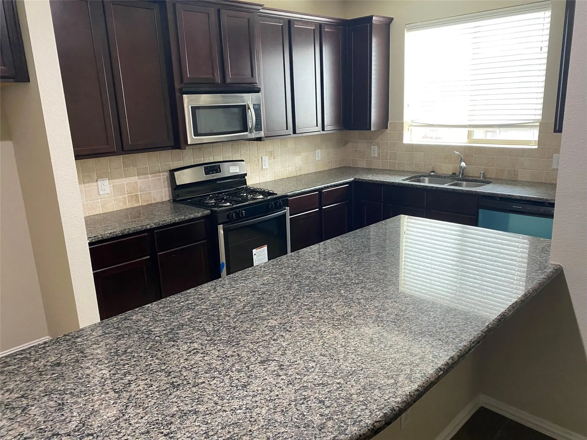 Kitchen featuring dark wood cabinetry, granite countertops, tile backsplash, stainless steel appliances, and a double basin sink with a window