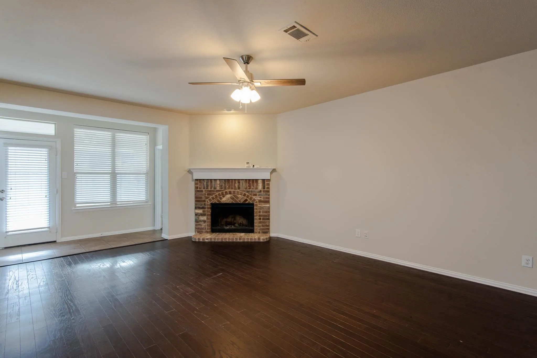 Spacious living area featuring dark wood-finish flooring, a brick fireplace with a white mantel, and a ceiling fan with integrated lighting