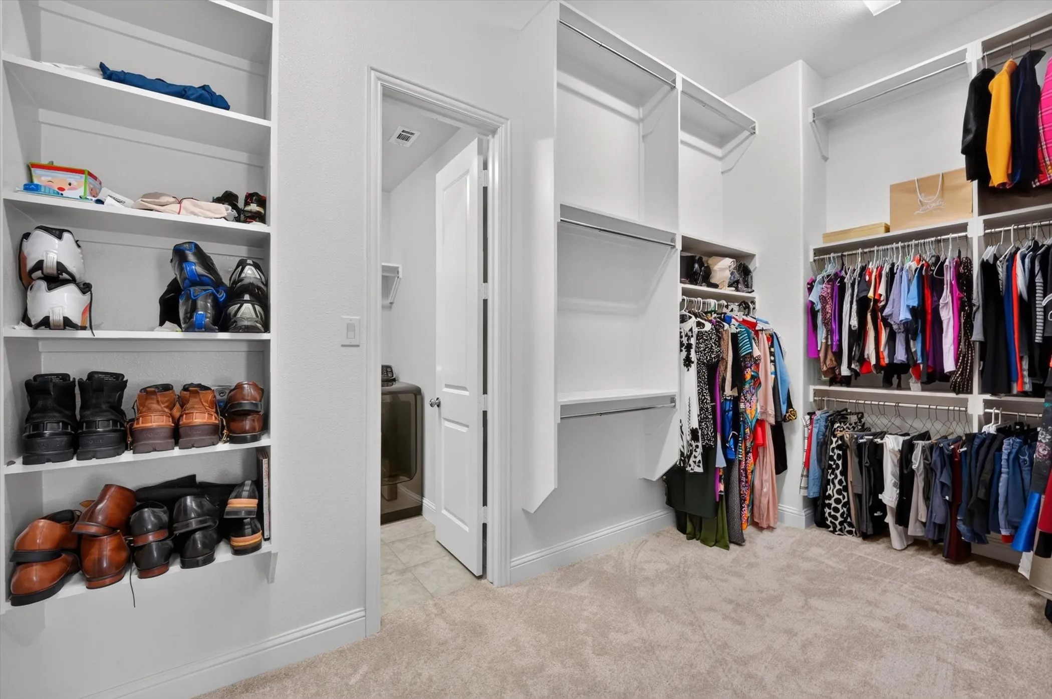 Spacious walk-in closet featuring light-toned carpet, white built-in shelving, multiple hanging rods, and an interior doorway with a white paneled door