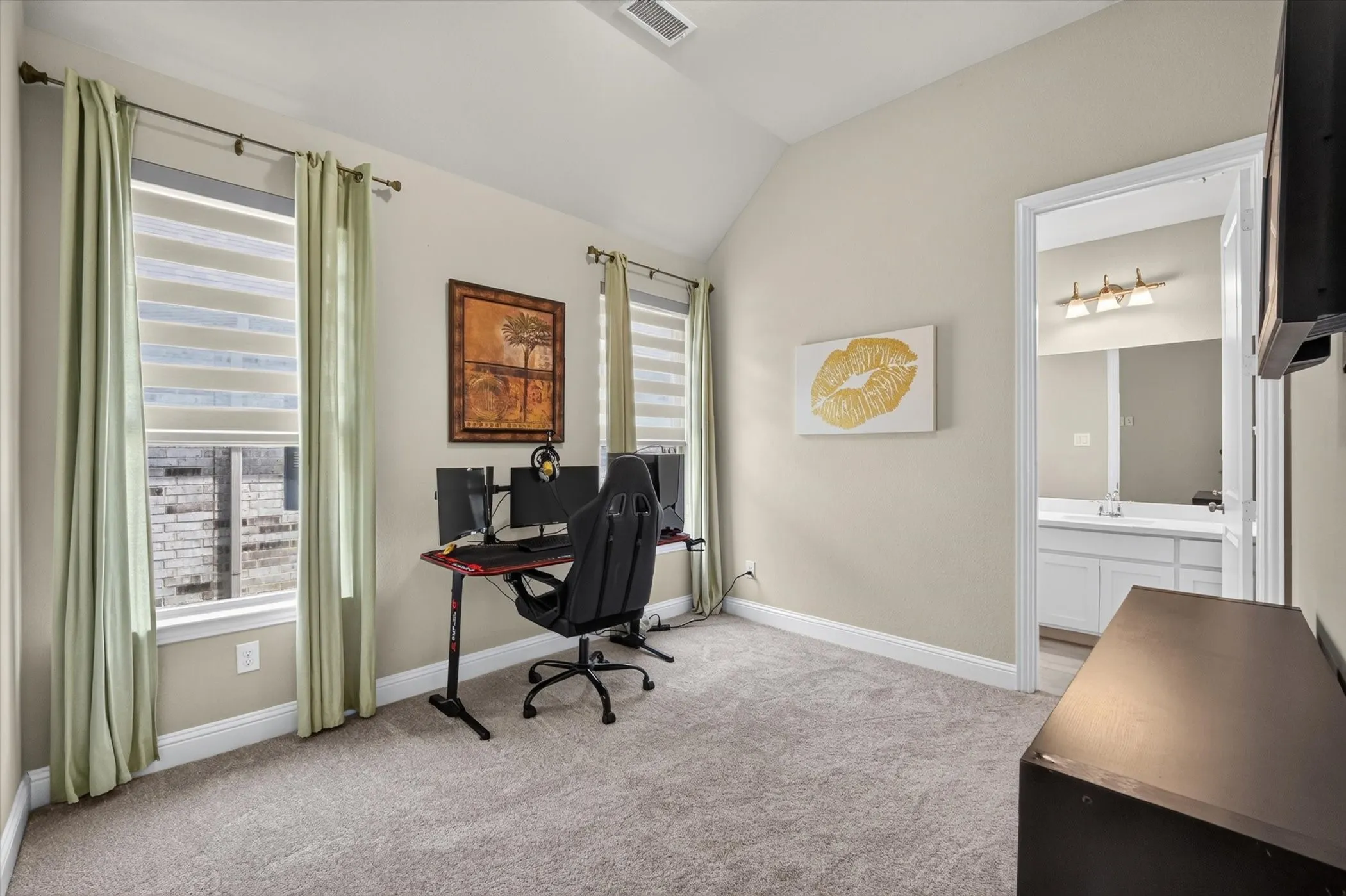 Carpeted interior room featuring two windows with horizontal blinds, a vaulted ceiling, and an entryway to a bathroom with a vanity and mirror