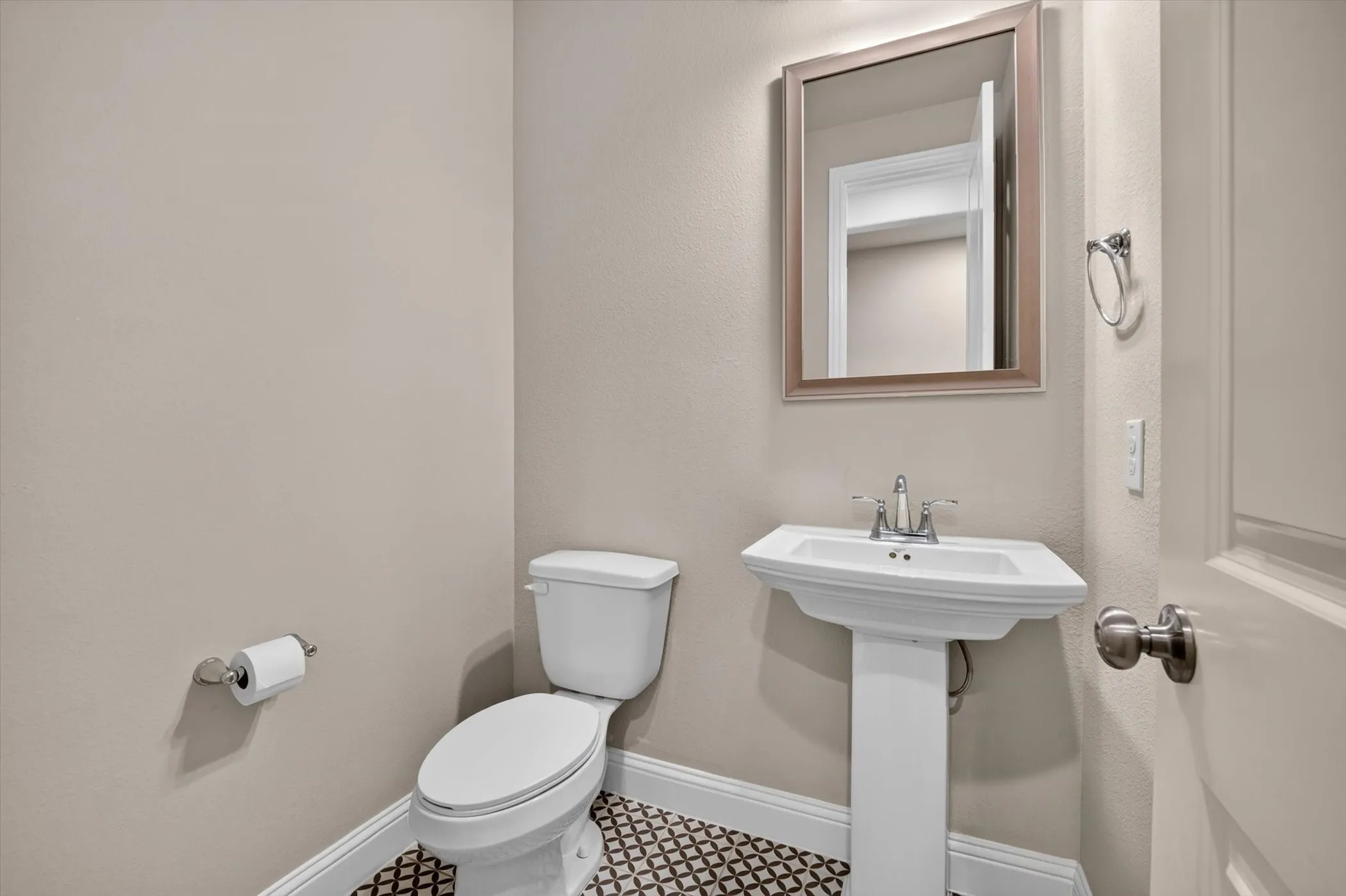 Pedestal sink with chrome hardware and a framed mirror above, complemented by a patterned floor tile, white baseboards, and neutral wall tones