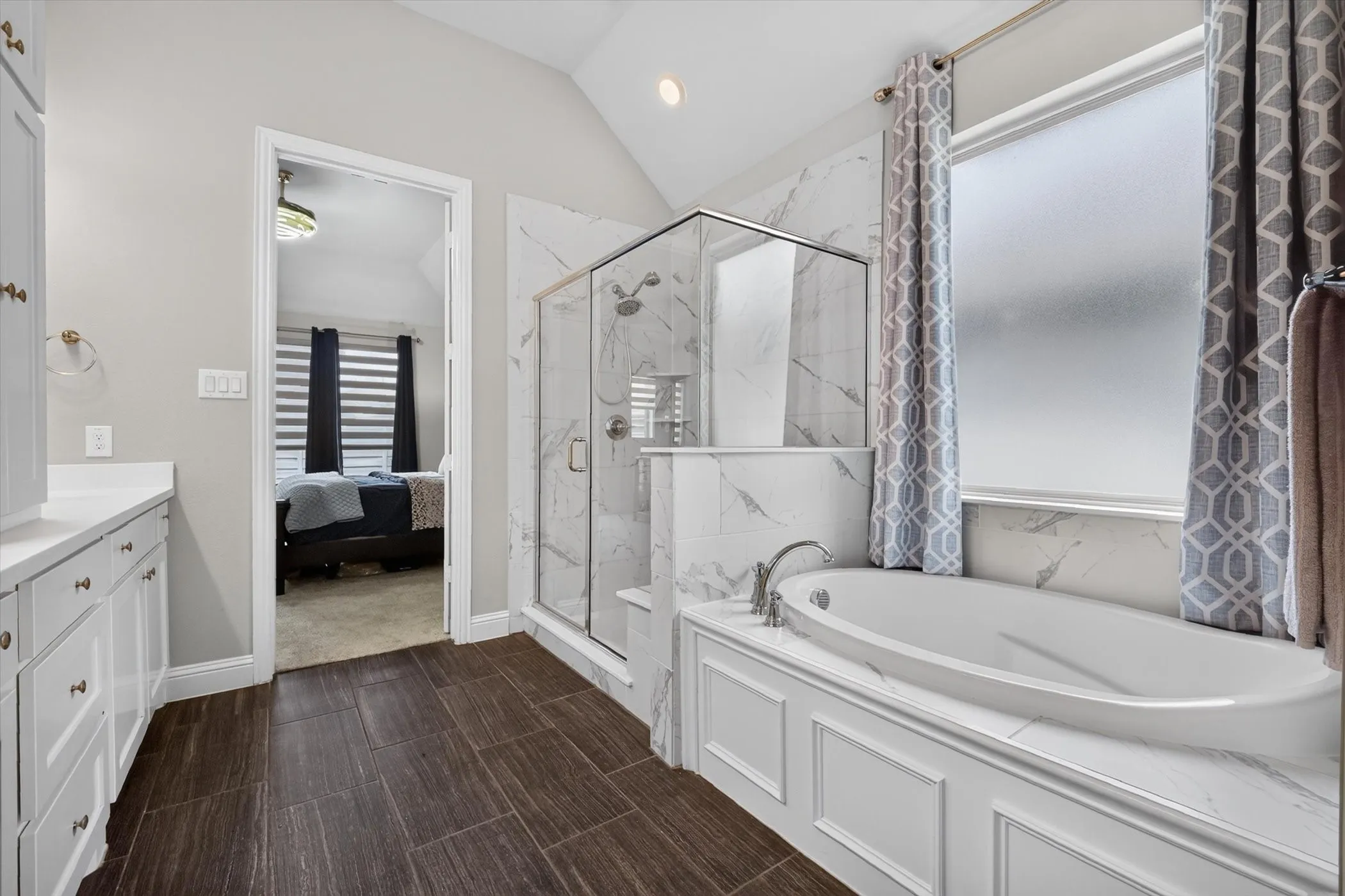Bathroom featuring dark wood-finish flooring, a freestanding soaking tub with paneling detail, a glass-enclosed shower with marble-look tile, and a double vanity with white cabinetry and light countertops