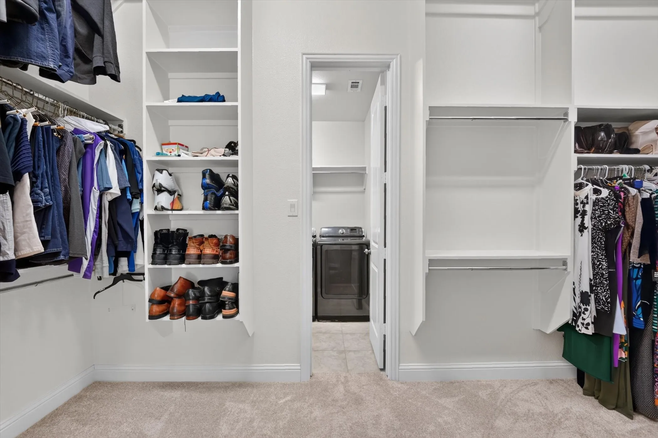 Walk-in closet with carpet flooring, built-in shelving, chrome hanging rods, and a doorway leading to an adjoining space featuring a built-in shelf and a dark-finished appliance