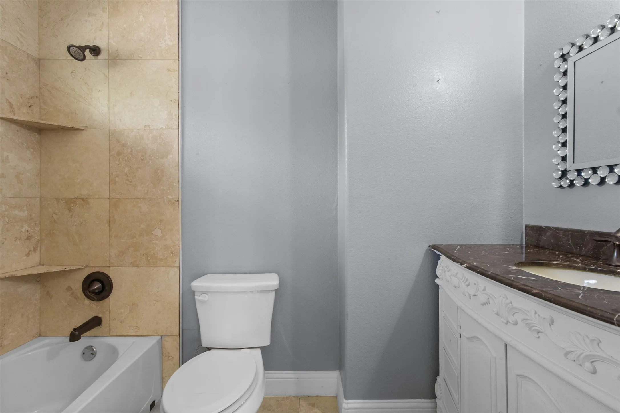 Bathroom featuring a built-in tub with a shower fixture, tile surround with integrated shelving, and a decorative vanity with an undermount sink and dark countertop